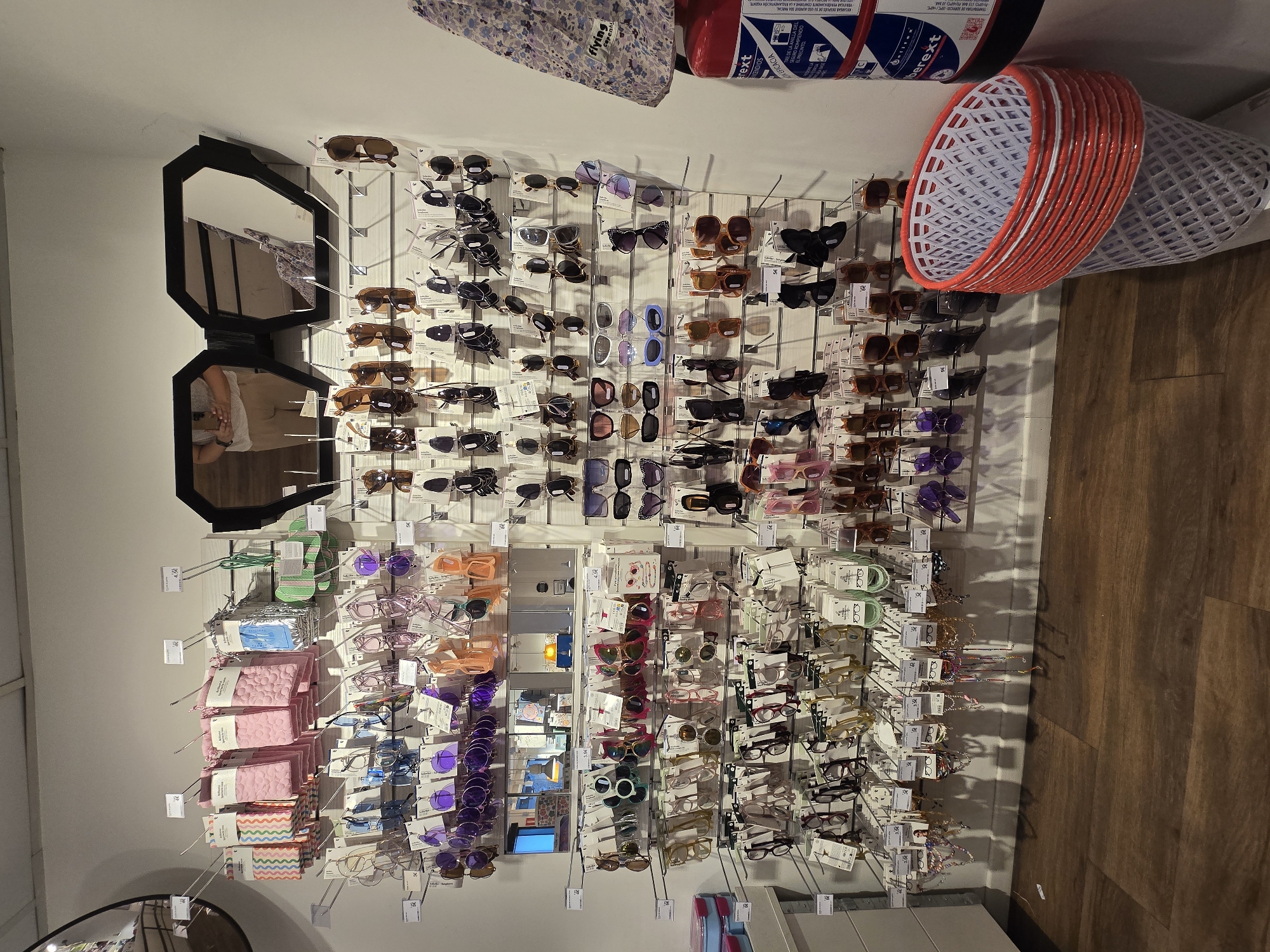 The image depicts a retail store display featuring an assortment of sunglasses and various accessories. The products are neatly organized on metal racks mounted on a white pegboard wall. Multiple rows of sunglasses, in a range of colors and styles, are displayed, each with its price tag. Above the sunglasses, there are shelves holding items such as patterned pouches, pink cloths, and what appear to be small cactus-shaped items. A large, geometric-shaped mirror with dark framing is mounted on the wall, reflecting a person holding a smartphone and the store interior. To the right of the display, a red fire extinguisher with a white label is visible, along with a white plastic basket with red trim. The floor is made of dark brown wood planks. The store appears to be an indoor commercial space, possibly a boutique or a department store. The scene is static and well-lit, focusing on the merchandise. There is no indication of the specific location within the city of Jaén, Spain, as no landmarks or city-specific identifiers are present in the image.