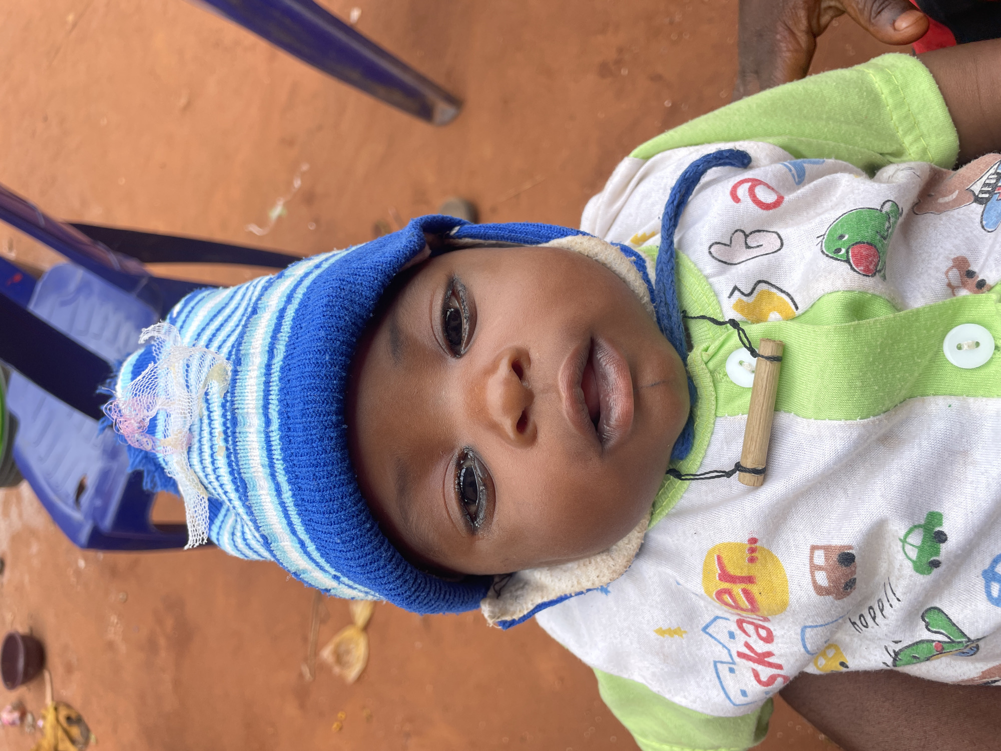 A baby wearing a blue and white striped hat and a white onesie with cartoon illustrations is lying down on a brown surface. A blue plastic chair is visible to the left. The location is Ogbe, Nigeria.