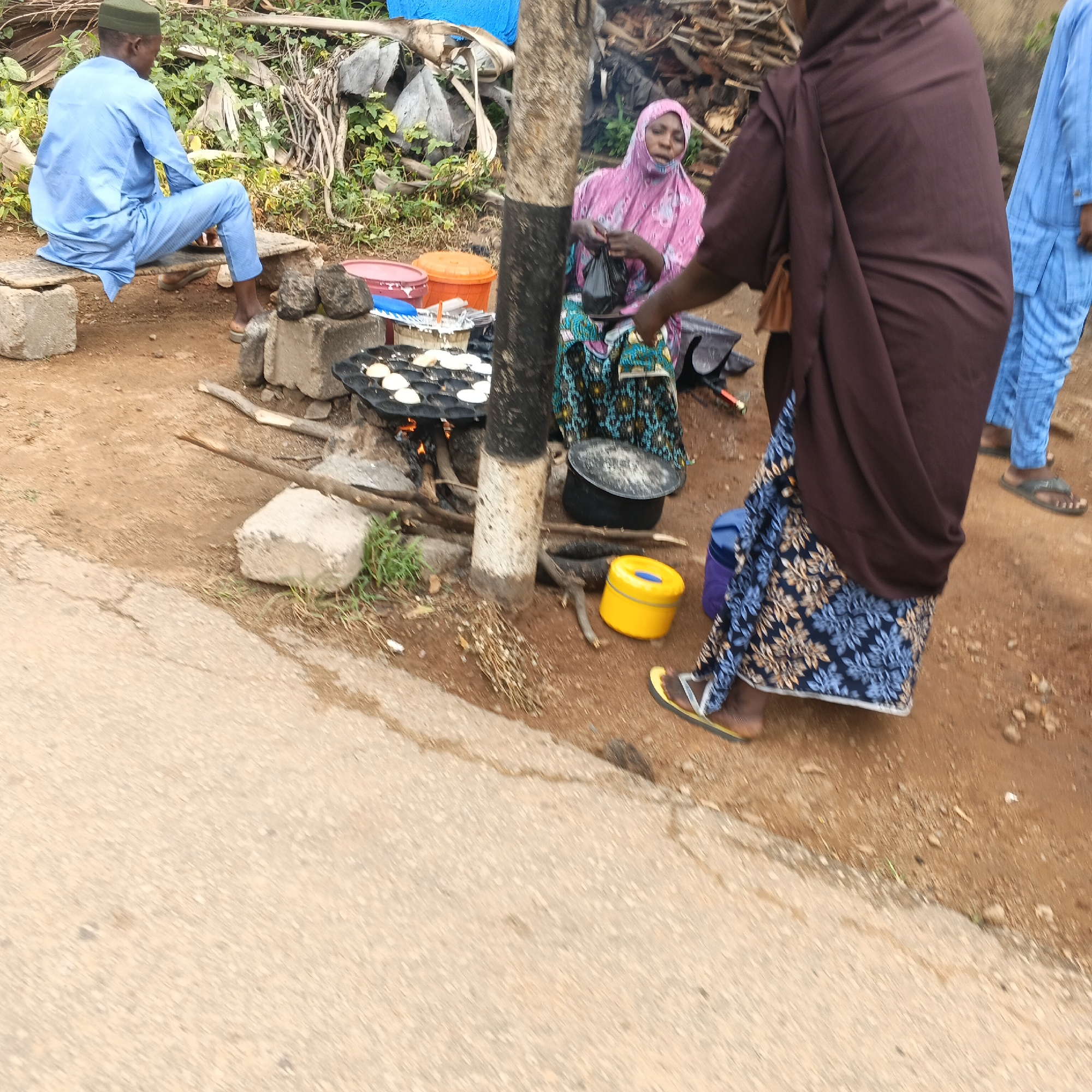 This image depicts a street scene in Unguwar Rimi, Nigeria, focusing on a food vending operation. In the foreground, a person in a blue outfit sits on a low bench made of concrete blocks and a wooden plank, facing away from the camera. To the right, a woman in a pink hijab and patterned green and blue skirt is actively preparing food. She holds a black plastic bag and appears to be interacting with the cooking setup. Next to her, another person, partially visible, wears a dark brown hijab and a long blue patterned skirt, standing and possibly observing or assisting. In the background, a person in a bright blue outfit, likely also a vendor or customer, is visible. The cooking area features a flat, circular griddle with multiple indentations, where small, light-colored food items are being cooked over an open fire fueled by wood. Several containers, including a bright yellow one with a blue lid, are present around the food preparation area. The ground is a mix of dirt and a paved surface, suggesting a roadside location. Vegetation and stacked wood are visible in the background, contributing to the rustic environment.