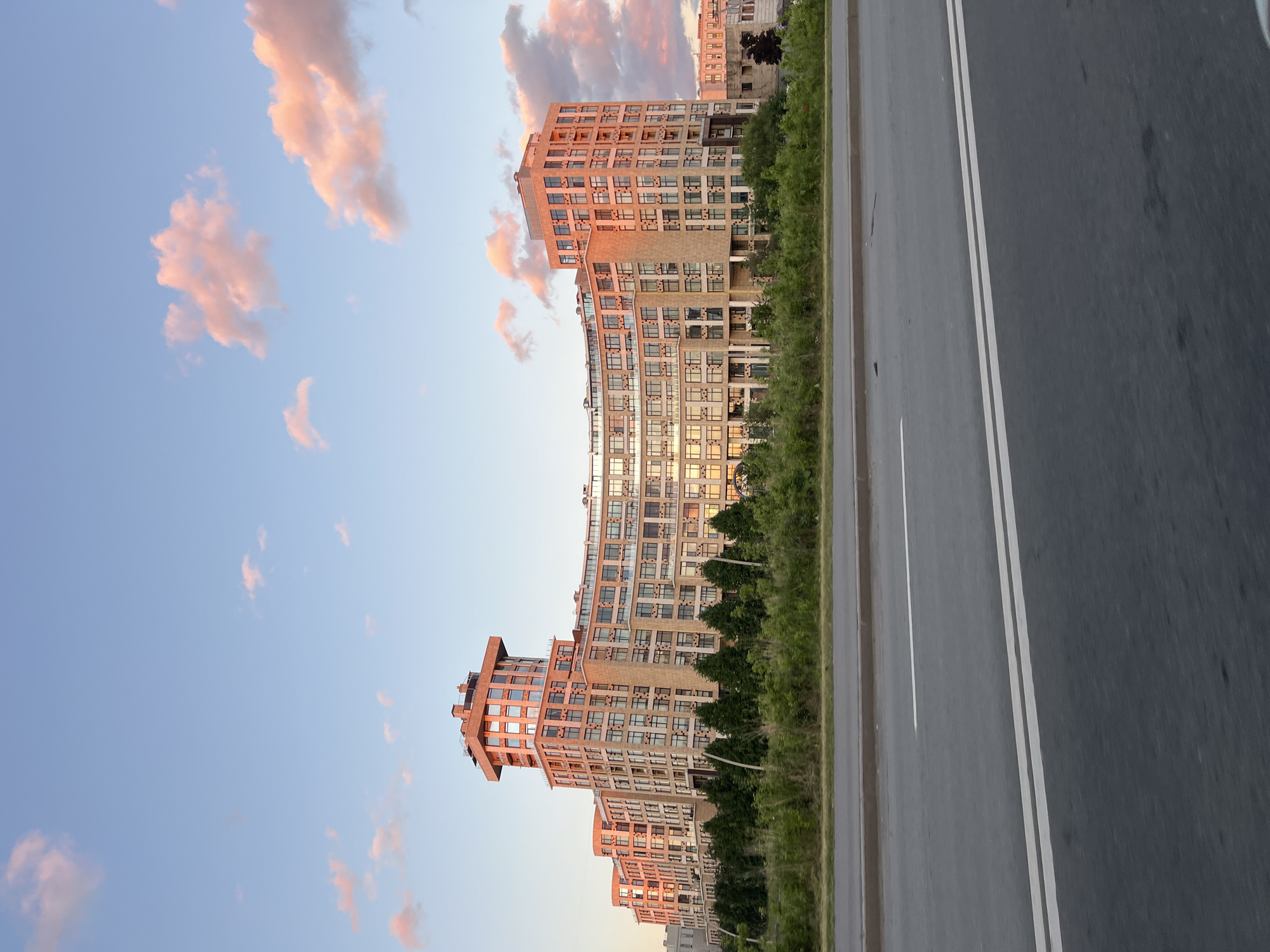 A wide shot captures a large, modern, beige apartment building with a curved facade, situated in Saint Petersburg, Russia, under a pale blue sky with scattered pink clouds illuminated by the setting sun. The building features numerous windows with dark frames, some reflecting the sky. A central, taller section of the building has a distinctive, blocky top. In front of the building, a line of dark green trees and bushes creates a natural barrier. A multi-lane asphalt road with white markings runs across the foreground of the image, suggesting the perspective is from a moving vehicle or roadside. The overall scene depicts an urban environment at dusk, characterized by its contemporary architecture and natural elements bathed in warm, golden light.