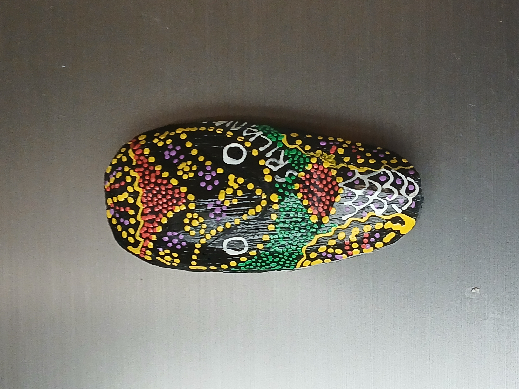 A painted rock rests on a metallic surface. The rock is black with multicolored dots (red, yellow, purple, green) and white lines that appear to form a snake-like image. White text is also visible on the rock. The background is a plain, light-colored, possibly brushed metal surface. The setting or location is indiscernible from the image. Bengaluru, India cannot be inferred from this image.