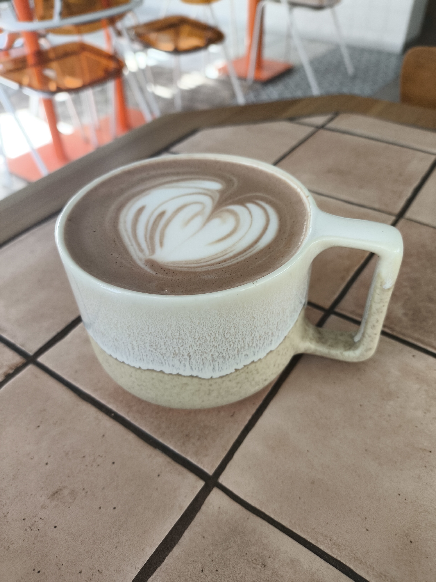 The image depicts a mug of hot chocolate with latte art on a tiled surface. The mug is two-toned, with a cream-colored upper portion and a beige lower portion. The latte art is a heart-shaped design created with white foam. The tiled surface is a terracotta color with dark grout lines. In the background, there are orange chairs and tables, suggesting the setting is a cafe or restaurant. The location is Unknown, Unknown.