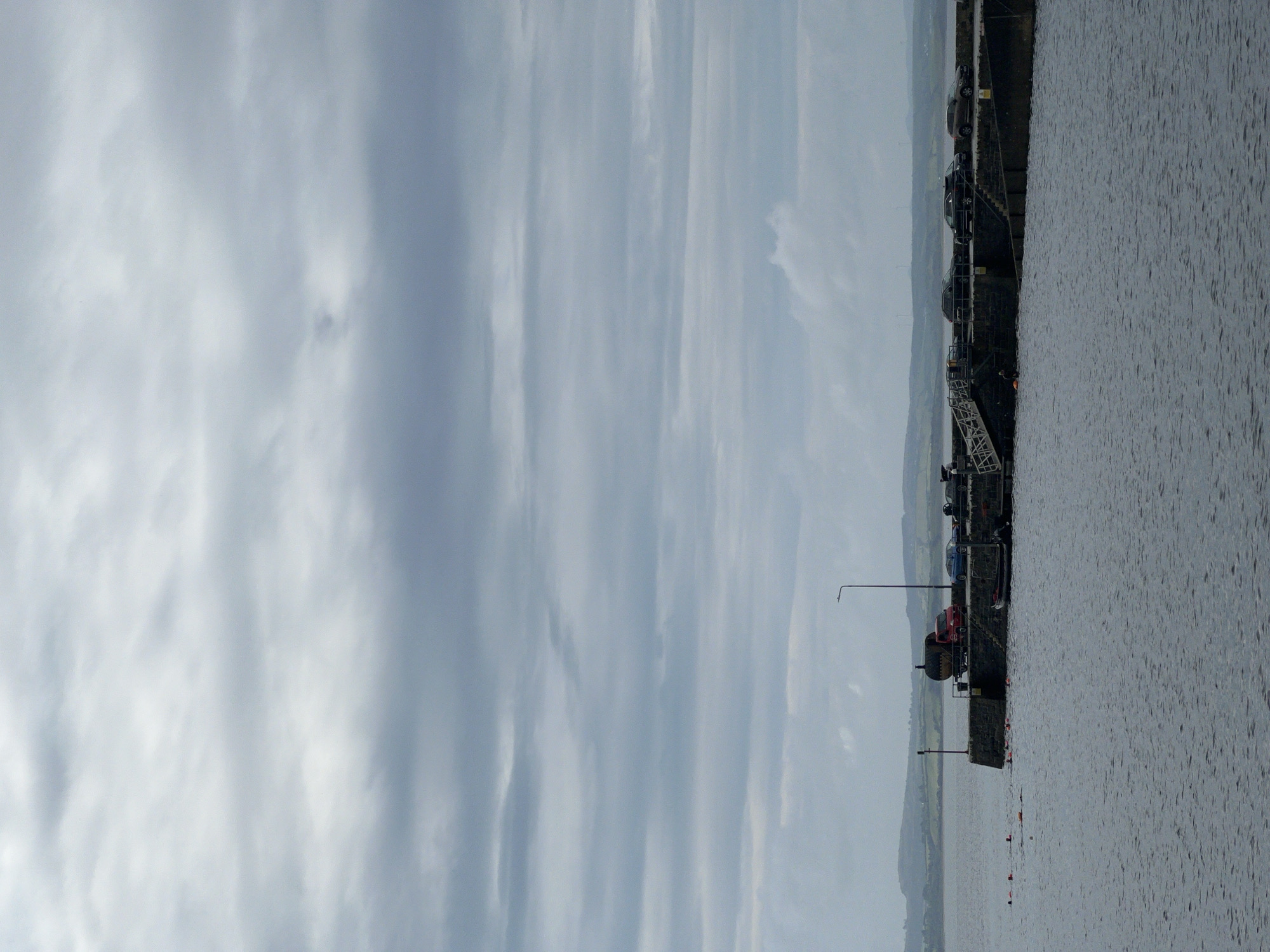 The image depicts a cloudy day over a body of water with a stone pier in the middle ground. The pier is constructed of rough-hewn stones and has several vehicles parked on it, including a red truck carrying a large dark cylinder, a blue car, and two darker colored SUVs. There are also a few people visible on the pier, and a smaller boat is docked. In the foreground, several orange buoys mark an area in the water where a group of people are swimming or training. The background shows a hazy coastline with rolling hills under a dramatic, overcast sky. The overall impression is one of a working harbor or waterfront area on a cool, gray day.