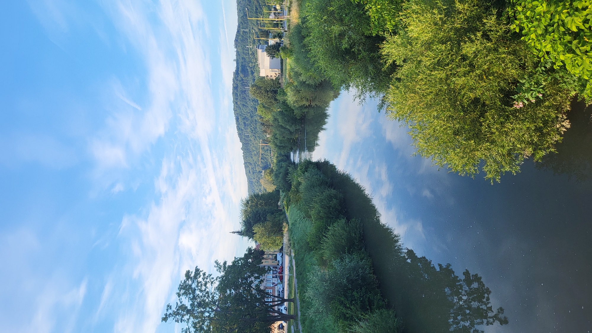The image depicts a serene river flowing through a verdant landscape, likely on the outskirts of Prešov, Slovakia, given the presence of a gently rolling hill in the background and urban elements to the left. The river, a dark, reflective body of water, is flanked by lush, dense vegetation, predominantly trees and bushes, creating a natural corridor. The water's surface mirrors the bright blue sky, which is adorned with wispy white clouds, suggesting fair weather.
On the left bank, a paved pathway runs parallel to the river, bordered by a grassy embankment. Beyond the grass, a row of trees stands, casting shadows on the pathway. Behind the trees, a street is visible with parked cars and buildings, indicating the proximity of a residential or commercial area. A lamppost is also present.
The right bank of the river is also covered in thick green foliage. In the distance, across the river and hills, a few modern buildings and construction cranes are visible, hinting at development within or near the city. The overall scene is tranquil and bathed in natural light, likely taken in the morning or late afternoon due to the soft illumination and the shadows. No people are visible in the image.