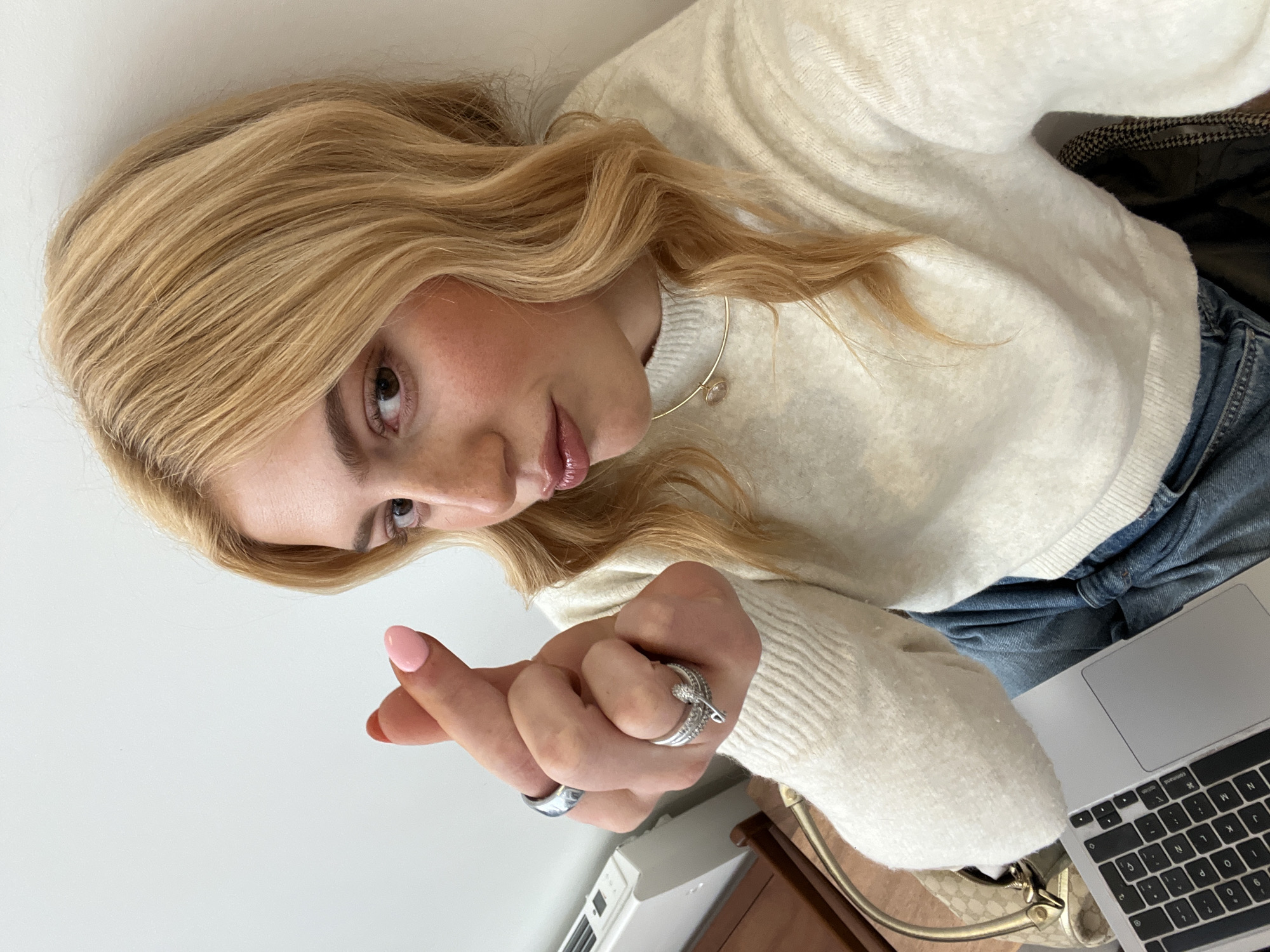 A young female, characterized by blonde, wavy hair and brown eyes, is depicted in a close-up, chest-up indoor setting in Paris, France. She wears a cream-colored knitted sweater and blue jeans. A gold-toned choker necklace with a clear circular pendant adorns her neck. On her left hand, she displays several silver-toned rings: a wide band on the index finger, and two rings on the ring finger—one plain and one ornate with small stones forming a serpentine design. Her left index fingernail is painted red, and her left thumb nail is pink. She makes a "finger heart" gesture with her left hand, crossing her thumb and index finger, and maintains a direct gaze towards the camera with a slight pout. The background features a plain white wall. To the left, a white wall-mounted radiator and a dark wooden surface are visible. In the lower left foreground, a silver-colored laptop and a beige monogrammed bag with gold-toned hardware are partially visible. A section of dark, patterned fabric, possibly upholstery, is visible in the lower right.