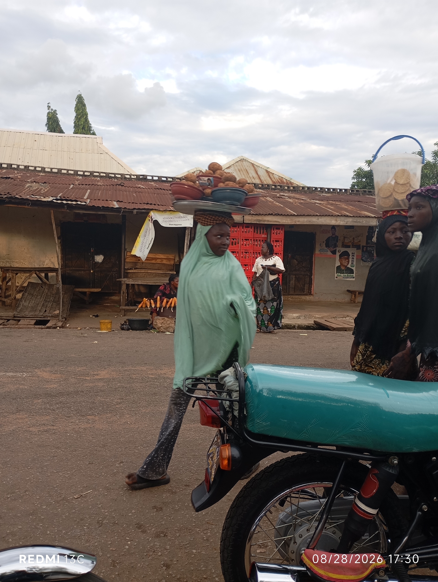 In the city of Shendam, Nigeria, a street scene unfolds with several individuals engaged in commerce. In the foreground, a young girl, draped in a light green hijab and patterned leggings, walks with a tray of round fruits, possibly coconuts, balanced on her head. She wears simple brown sandals. Behind her, a turquoise motorcycle seat is prominently displayed. Further back, the environment suggests a marketplace or roadside stalls. Two women, one in a dark hijab and patterned fabric, and another in a lighter-colored garment, stand near a stall. A third woman in the background wears a white top and a patterned skirt, carrying a dark bag. Stalls with corrugated metal roofs line the street, some displaying goods. One stall has a prominent poster featuring a man's face with text, likely a political advertisement. Another stall appears to be selling corn on the cob, with several skewers of corn displayed on a wooden block. The ground is paved with asphalt. The sky is overcast, with a mix of clouds. A date and time stamp, "08/28/2026 17:30", is visible in the lower right corner.