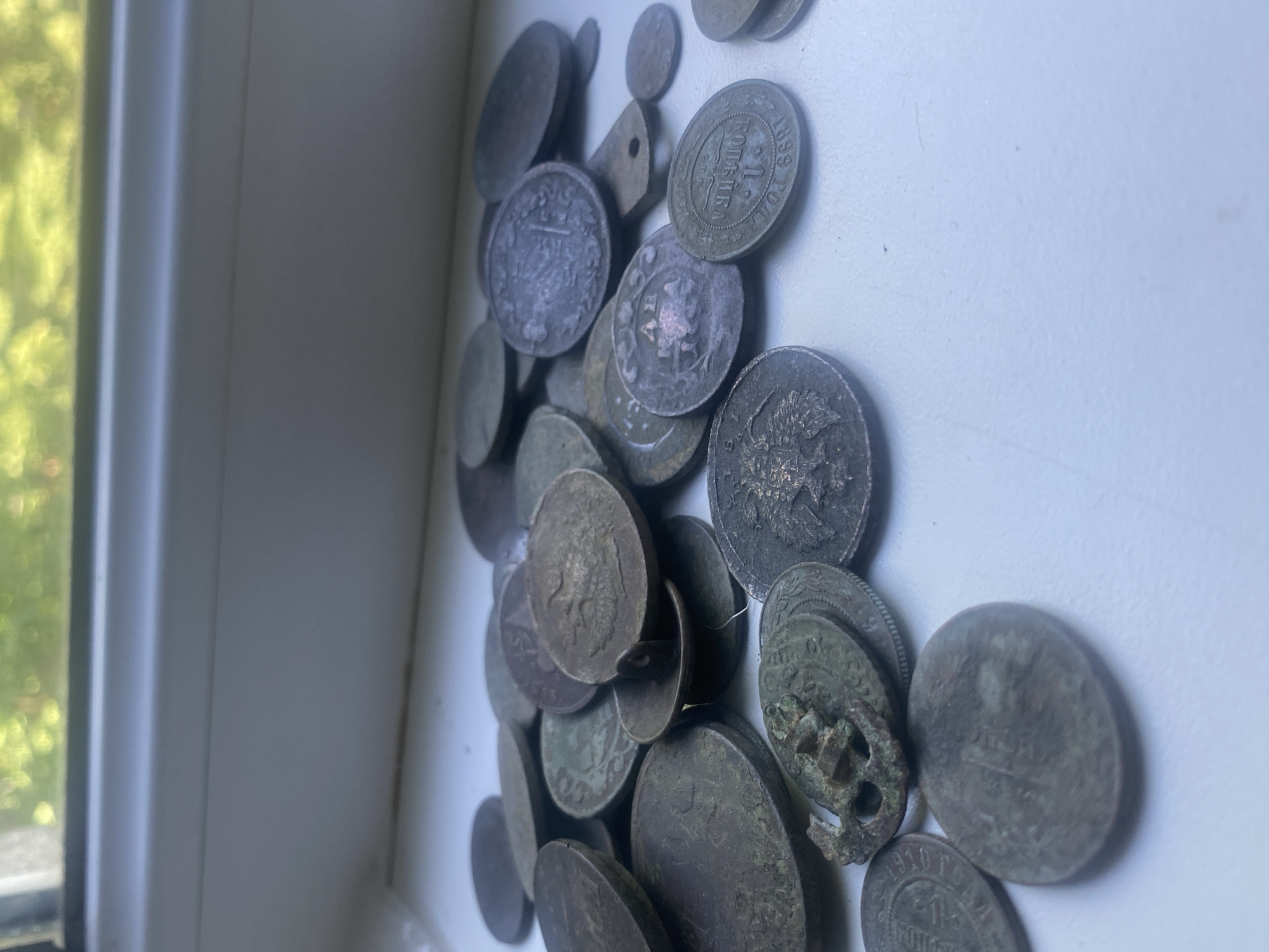 A collection of old coins is scattered on a white surface, possibly a windowsill, with a blurred background showing green foliage outside a window. Some of the coins show details like engravings, dates, and possibly denominations, but most are tarnished. The coins are of varying sizes and metallic tones. There is no indication of people, actions, or specific interactions. The location is not definitively identifiable as Poltava, Ukraine based on the provided image alone.