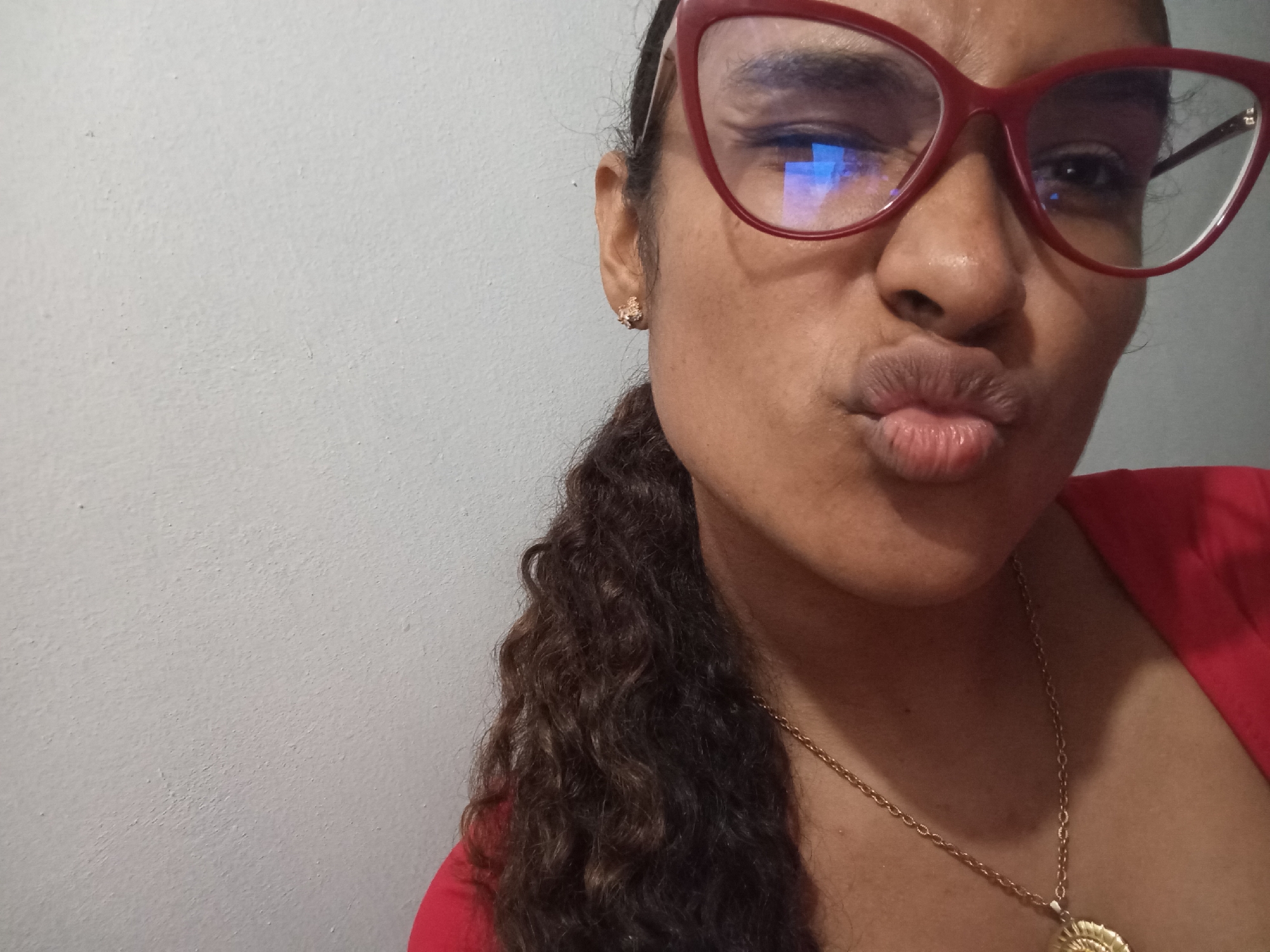 A medium close-up shot shows a person with brown curly hair pulled into a low ponytail on the right side of the frame. The person is wearing red-framed glasses and has on a gold necklace. They are pouting, with their lips pursed. The background is a plain white wall. There is no indication of the location being Juan Griego, Venezuela.