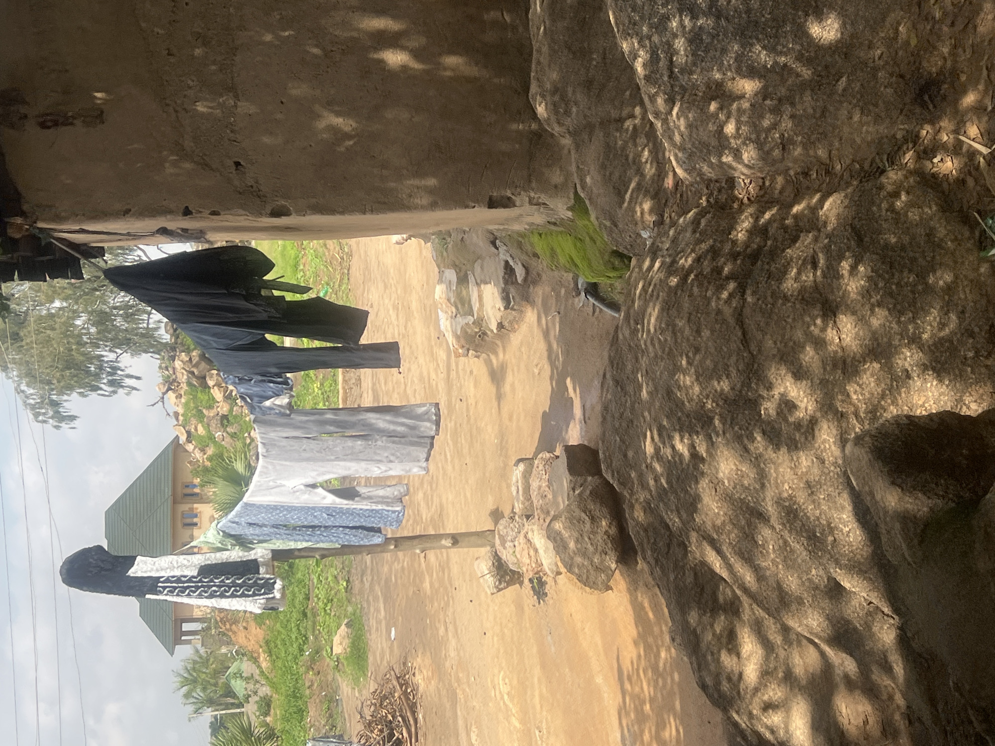 The image shows a scene in Gura Top, Nigeria, where laundry is being hung to dry. A black garment is draped over a clothesline, followed by several lighter-colored garments, including a patterned blue and white shirt and a grey shirt. The clothesline appears to be made of a thin rope or wire and is supported by a wooden post. In the background, a house with a green roof and beige walls is visible, surrounded by greenery and palm trees. Large, weathered rocks are a prominent feature of the landscape, casting shadows on the ground, which suggests it is daytime. The ground is a dusty, sandy color, with some scattered dry leaves and twigs. The overall setting appears to be outdoors, possibly in a rural or semi-rural area. The lighting suggests it is a sunny day. No people are visible in the image.