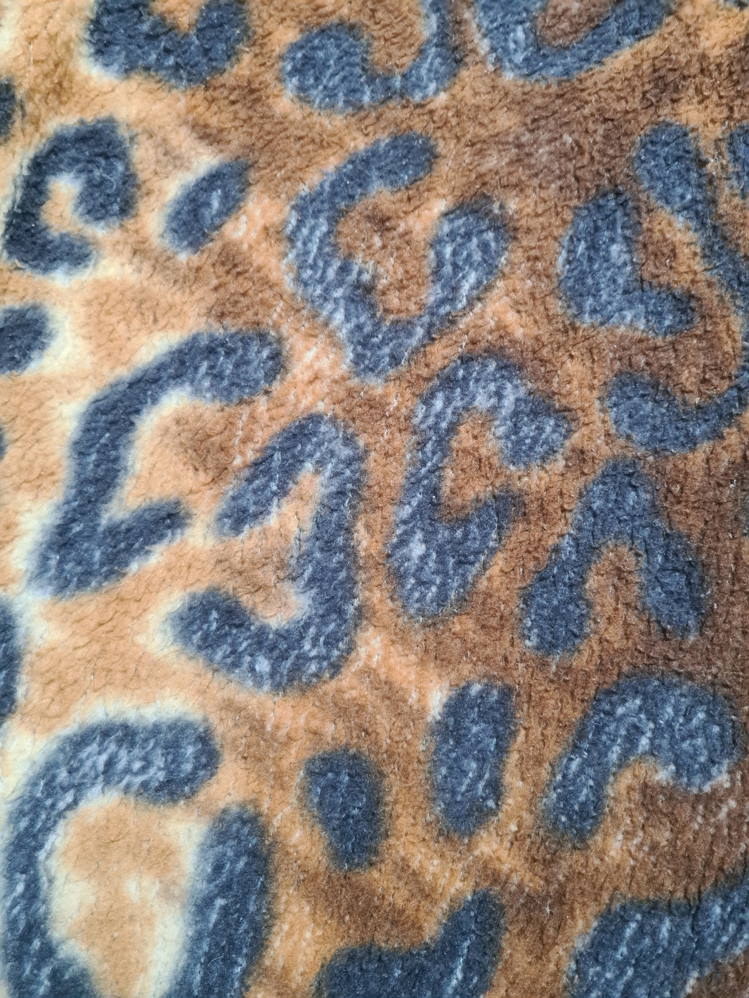 The image is a close-up, overhead shot of a leopard print fabric. The print features irregular spots and splotches of dark grey and brown on a light tan and cream background. The texture of the fabric appears soft and possibly plush, with visible fibers. There are no people or identifiable objects other than the fabric itself. There is no information in the image to suggest a location in the city of Rijeka, Croatia.