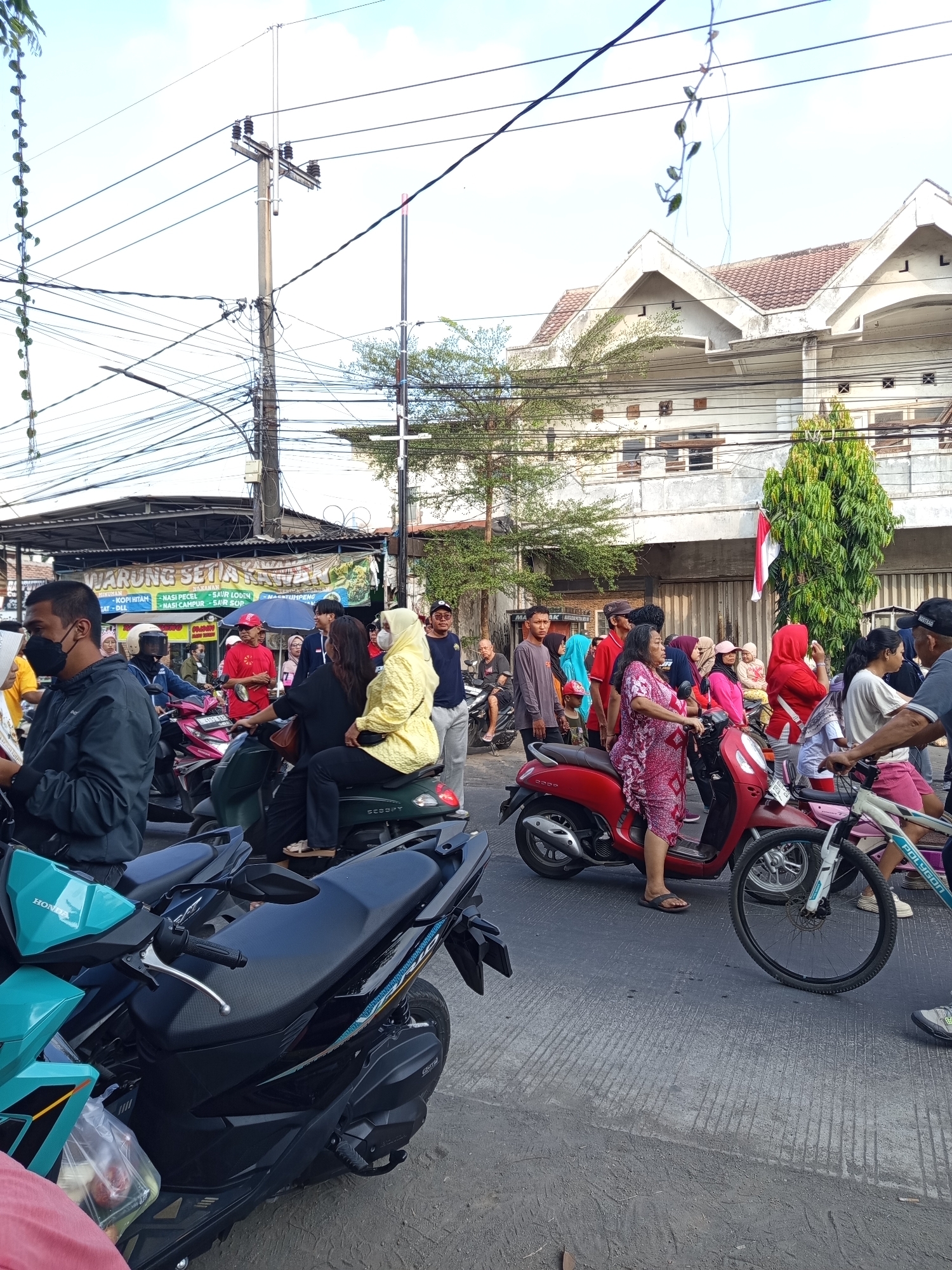 The image shows a street scene in Suko, Indonesia, with a crowd of people and several motorcycles. A banner for "WARUNG SETIA KAWAN" is visible, indicating a food stall or restaurant. People are seen on and around motorcycles, some in motion, suggesting a busy street or an event. The environment appears to be urban, with buildings and overhead power lines. An Indonesian flag is draped on a building. The overall atmosphere seems to be lively with a significant number of individuals gathered.