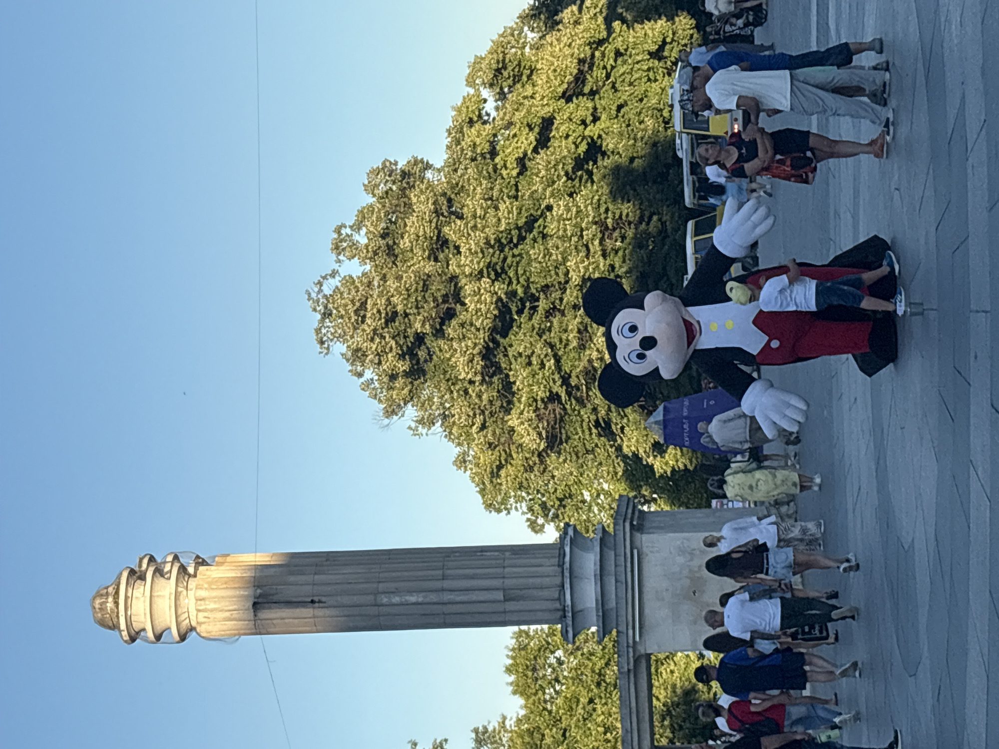 In a public square, a costumed Mickey Mouse character stands in the foreground, facing right with arms outstretched. A young boy wearing a yellow cap and blue shorts stands next to Mickey Mouse, looking up at the character. Behind them, a crowd of people walks in various directions. A tall, fluted column with a weathered top stands to the left, with an archway visible behind it. Lush green trees with dense foliage fill the background. Several yellow vehicles, possibly tour buses, are parked in the distance. The scene is illuminated by a clear blue sky, suggesting it is daytime.