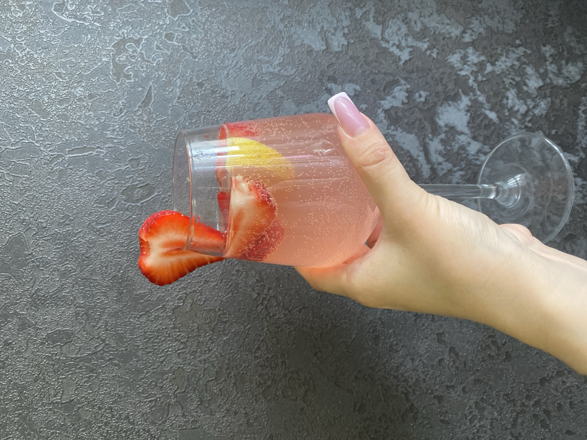 A woman's hand with long, manicured nails (French manicure) is holding a tilted champagne glass. The glass contains a pink, bubbly liquid, presumably a cocktail, garnished with slices of strawberry and lemon. One strawberry slice is partially outside the glass. The glass is resting against a textured, dark gray surface, possibly a countertop or wall. The image appears to be taken indoors. There is no verifiable information in the image regarding the city of Izhevsk, Russia.