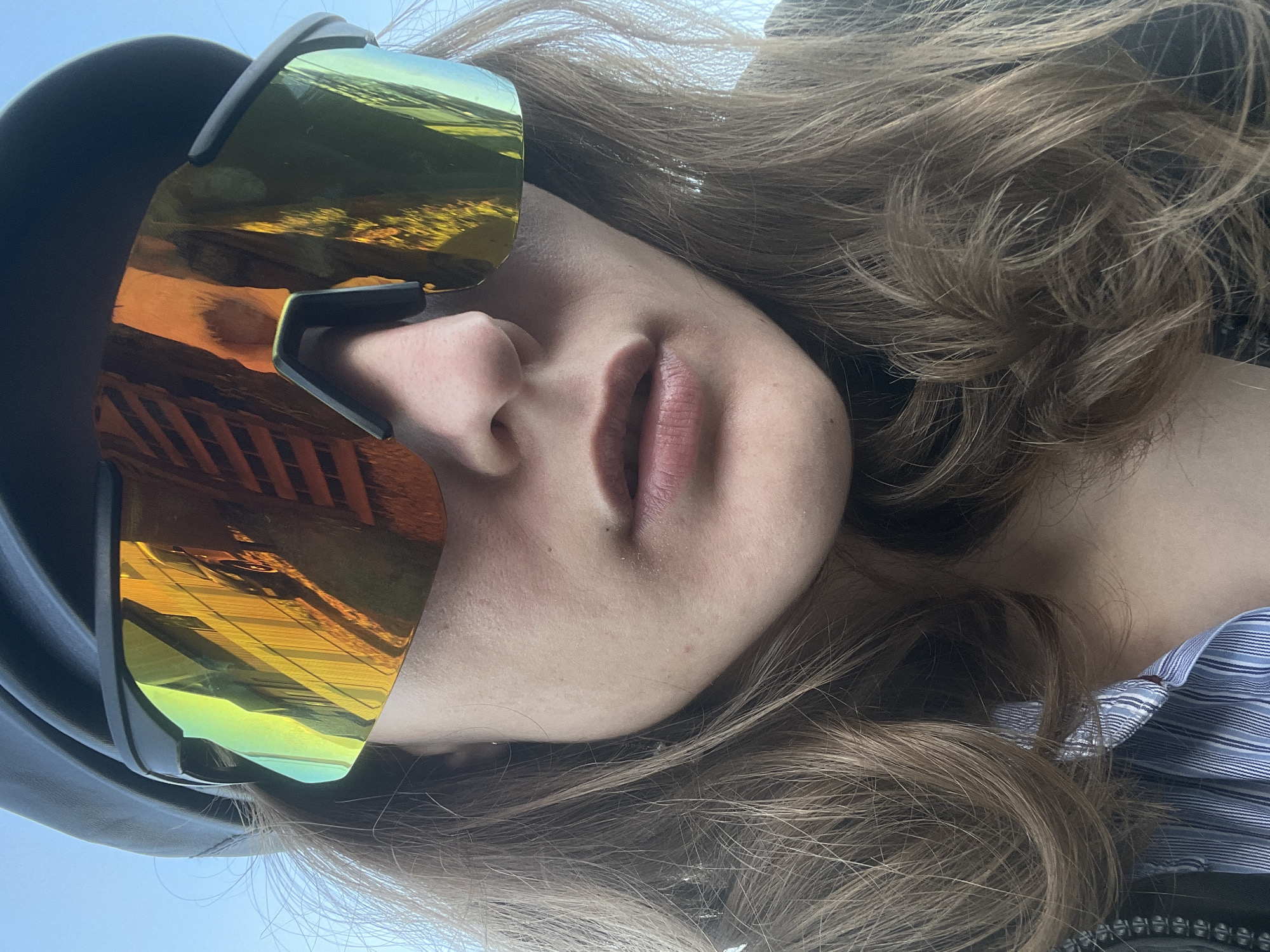 A close-up, rotated image shows a fair-skinned person wearing large, reflective gold-tinted sunglasses. Only their nose and mouth are fully visible beneath the glasses. The reflection in the lenses appears to show an outdoor urban scene with architectural details. The person's hair is light brown and wavy, falling around their face and shoulder. They appear to be wearing a black hat and a blue and white striped shirt. It is impossible to confirm that the image was taken in the city of Подшивалово, Russia.