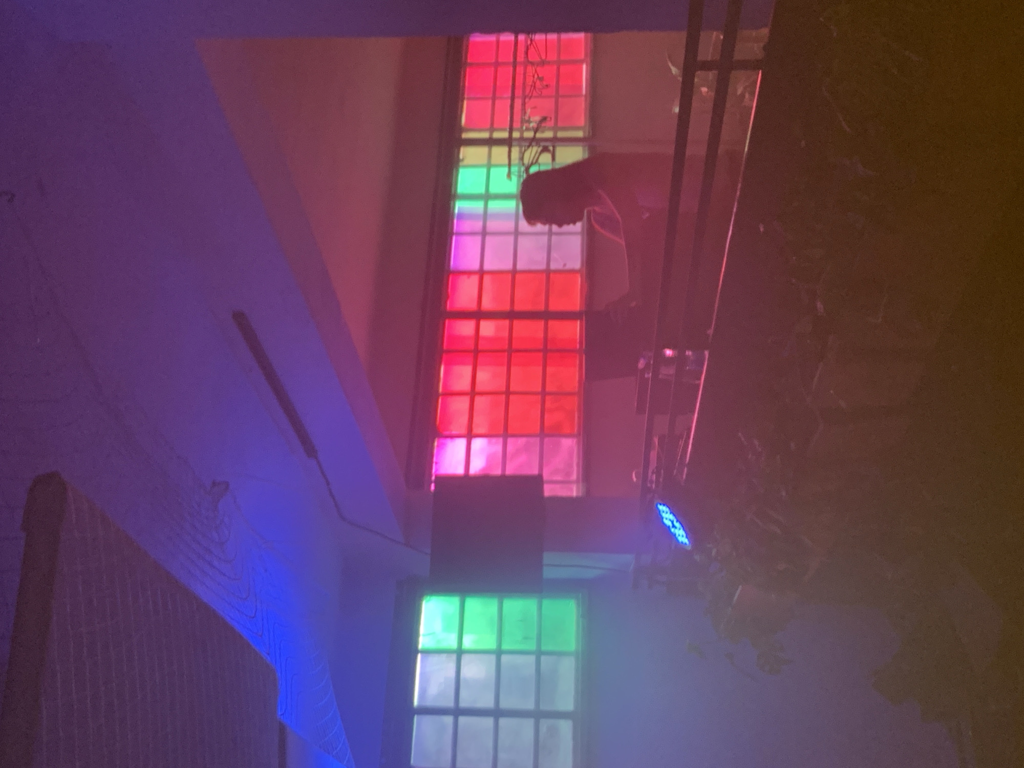The image appears to be taken indoors, looking towards two windows. The upper window is divided into a grid pattern and filled with colored panes of glass, predominantly red, pink, and green. The lower window is also divided into a grid pattern, with panes filled mostly with green. A person's silhouette is visible against the upper window. To the left, a textured wall is illuminated with blue light. To the right, an indiscernible object or surface is illuminated in yellow or amber light. The scene is in a space with purple and pink lighting. There is no visible indication of the location being Tel-Aviv, Occupied Palestine.