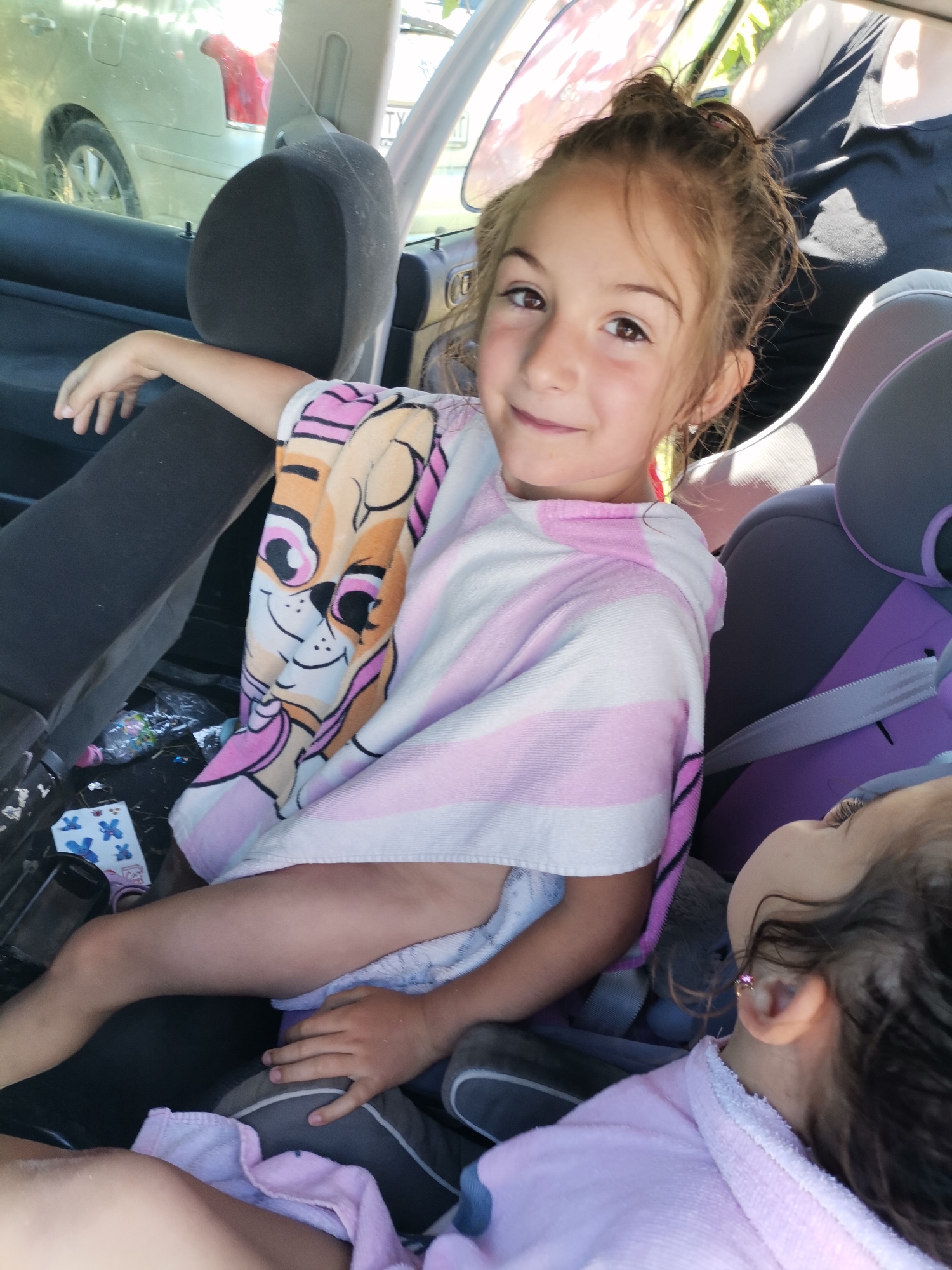 In the back seat of a car, two young girls are sitting, possibly on a trip. The girl in the foreground, on the left, is looking at the camera with a smile. She is wearing a pink and white striped hooded towel featuring a character from Paw Patrol. Her arm is resting on the back of the car seat. To her right, another young girl is visible, also wearing a pink hooded towel. Her face is partially obscured as she looks upwards. The car interior is visible, with car seats and a glimpse of the outside through the windows. The scene appears to be during the daytime, with bright sunlight suggesting fair weather. The location context is Kranevo, Bulgaria. There is no visible text in the image.