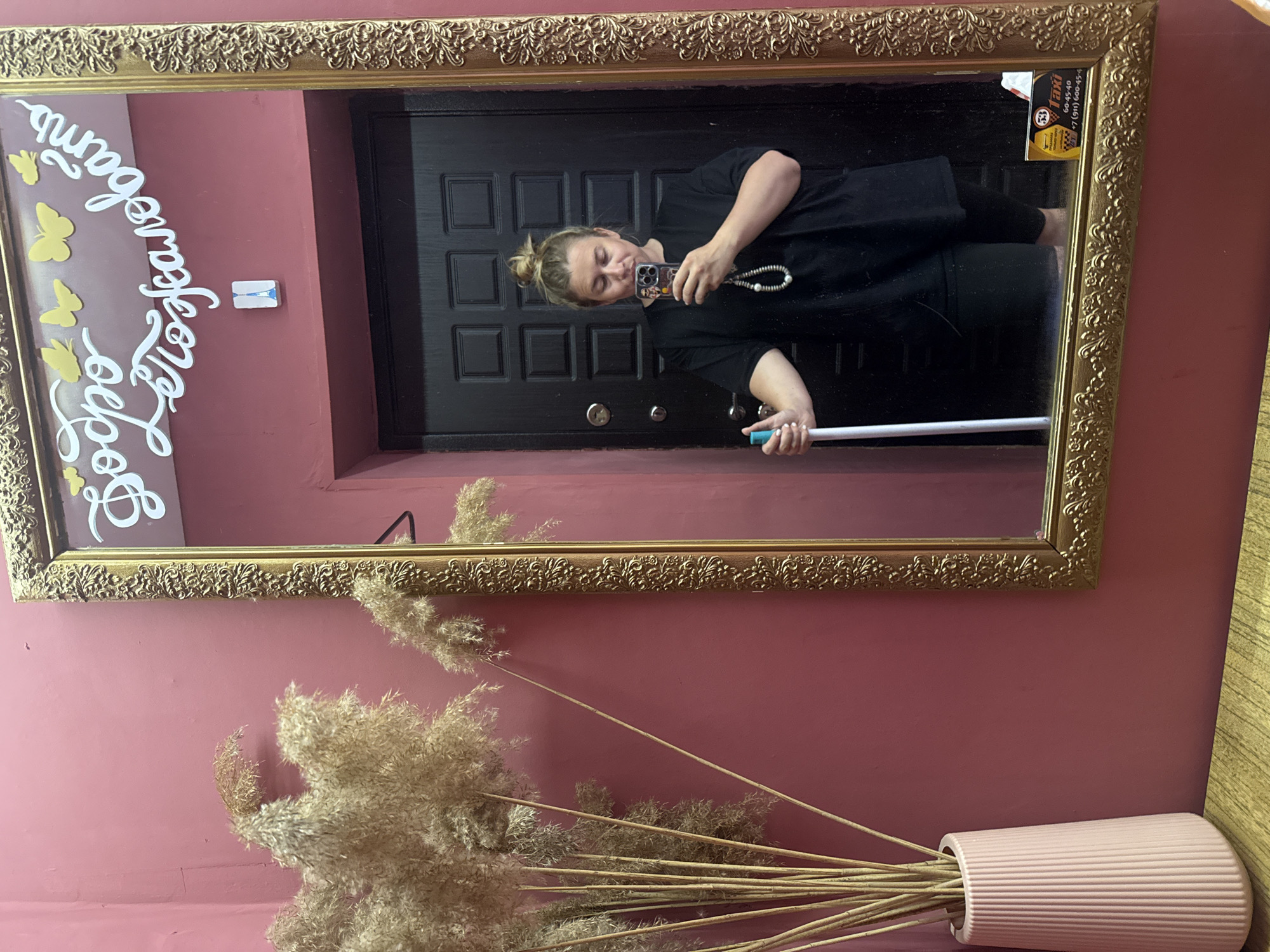 A woman is reflected in a large, ornate gold-framed mirror. She is standing in front of a dark, paneled door, holding a white mop handle in her left hand and a smartphone in her right hand, taking a selfie. She has blonde hair pulled up in a bun and is wearing a black t-shirt and dark shorts. A beaded strap hangs from her phone. The mirror is decorated with white lettering that reads "Добро Пожаловать" (Welcome) and several yellow butterfly cutouts. A small rectangular sticker depicting the Eiffel Tower is attached to the wall above the door. To the left of the mirror, a tall pink vase holds a large bunch of dried reeds. The wall behind the vase and mirror is painted a dusty rose color. A yellow and black taxi advertisement is visible on the lower right edge of the mirror. The scene appears to be indoors, possibly in an entryway or hallway. The city of Valday, Russia is mentioned as the location.