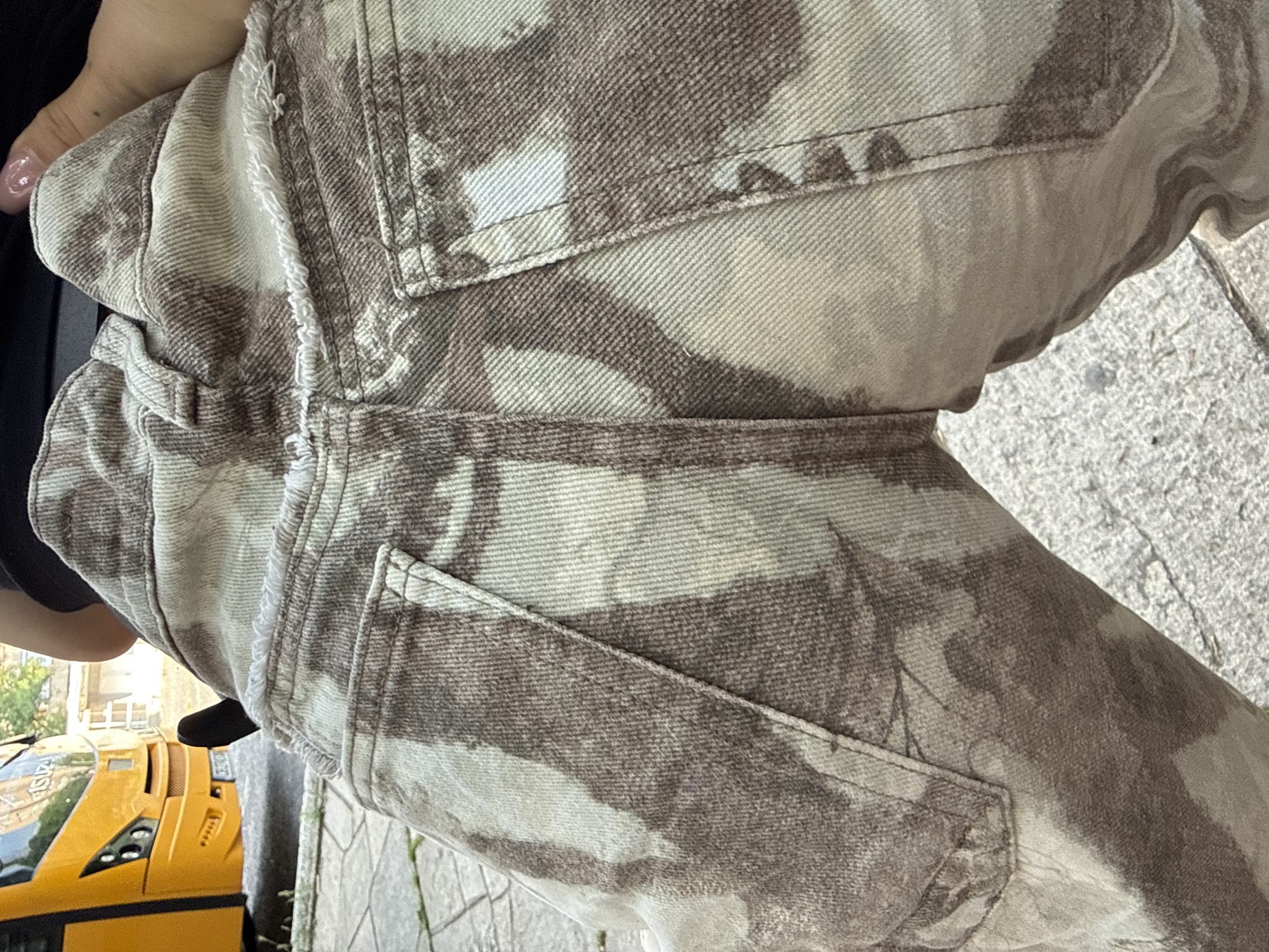 The image, set in Kardzhali, Bulgaria, presents an angled, close-up view of a person's lower body, specifically their right leg, dressed in patterned denim pants. The pants feature a distinct mottled or tie-dye pattern in varying shades of light brown, beige, and off-white, with a visible diagonal twill weave. Details like stitched back pockets, belt loops, and frayed, distressed edges along seams are clearly visible, contributing to a casual style.
In the upper left corner, a hand with manicured nails, painted a light pink or clear shade, is partially visible. The person appears to be standing or walking on a textured paved surface, likely a sidewalk or pedestrian area, made of concrete or stone blocks with visible cracks.
A bright yellow car is positioned to the left and slightly below the person. The car's design, with its distinctive headlights and vents, suggests a sports car. However, its proportions and the clear "ISO" lettering on its side lead to the impression that it might be a large-scale model, a toy, or a child's ride-on vehicle rather than a full-sized car. A partially visible license plate on the car's rear displays the numbers "3002" and the regional code "KA," consistent with Kardzhali, Bulgaria.
The scene is well-lit by bright daylight, suggesting good weather. Blurry indications of greenery and distant structures are present in the background. No specific activity beyond standing or movement is explicitly captured, and no emotions are discernible from the visible parts of the subject.