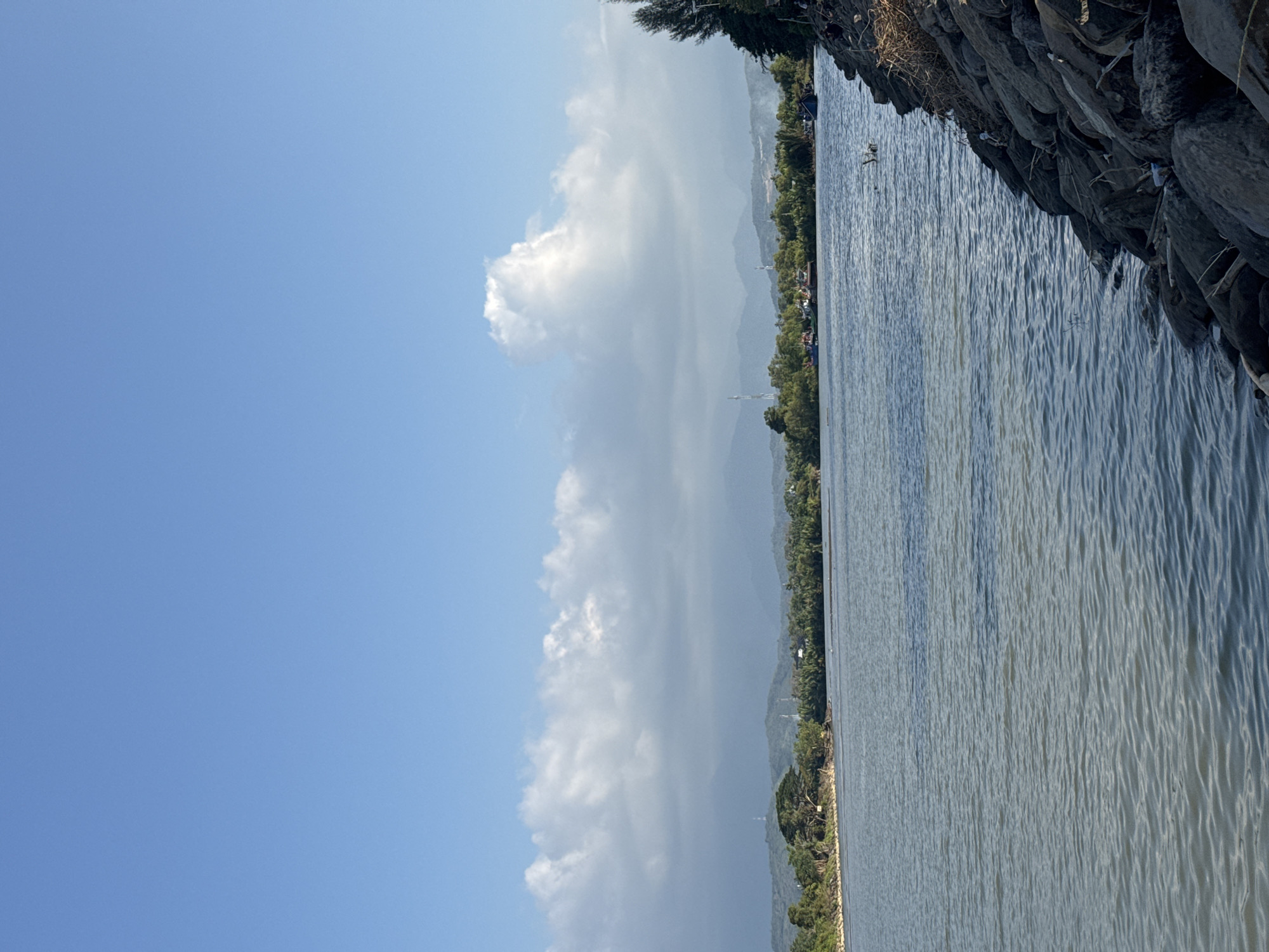 The image depicts a scenic view of the waterfront in Reudeuep, Indonesia. The sky is a clear, bright blue with scattered fluffy white clouds. In the foreground, a body of water, likely a river or bay, is visible, with gentle ripples on its surface. Along the right bank, a rocky embankment is present, with some dried vegetation and debris scattered among the stones. The left bank of the water features lush green trees and vegetation, with a few buildings and structures interspersed. In the distance, rolling hills and mountains form the backdrop, partially obscured by atmospheric haze. Several boats are anchored or moored in the water near the left bank, and a single, dark-colored boat is visible in the water on the right side. The overall scene conveys a sense of natural beauty and tranquility.