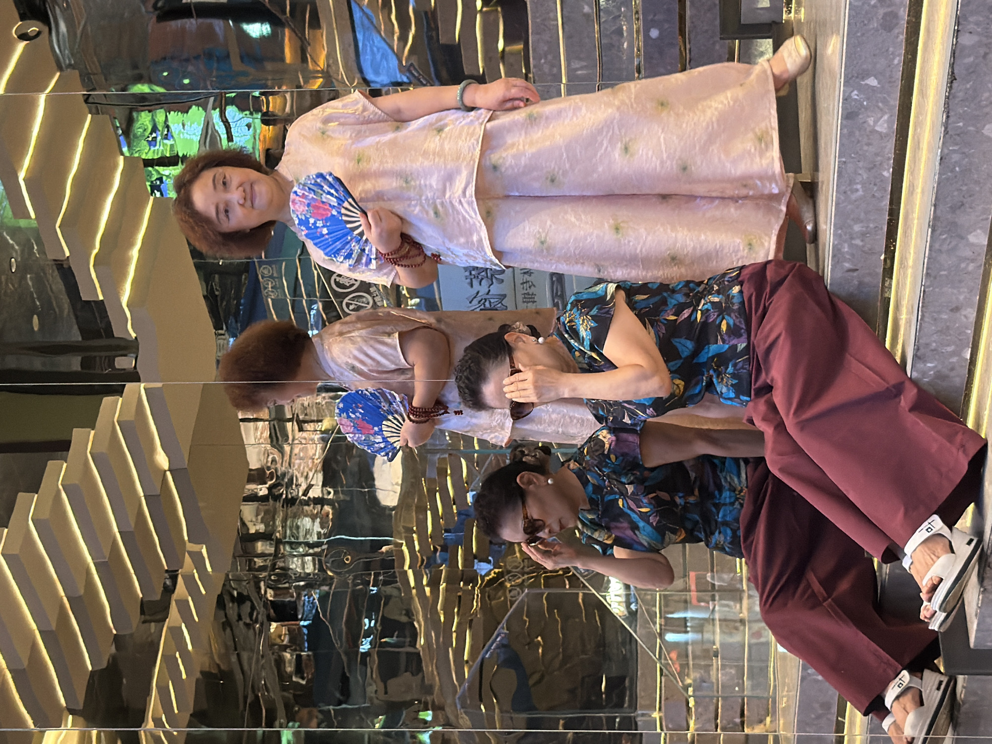 In Yuzhong District, China, four women are depicted in what appears to be a modern indoor setting with mirrored walls and illuminated tiered structures resembling stairs or shelves. Two women are seated on a step, each wearing a dark, floral-patterned top and burgundy wide-leg pants, with sunglasses and white sandals. The woman on the left has her hand raised to her sunglasses. The other two women are standing. The woman on the right, in the foreground, is wearing a light pink, patterned loose-fitting outfit and holding a blue and pink fan. She is also wearing a bracelet. Behind her, another woman, partially visible and seen from the side, is also wearing a similar light pink outfit and holding a blue fan. The background is dominated by the reflective surfaces, creating multiple reflections of the subjects and the environment.