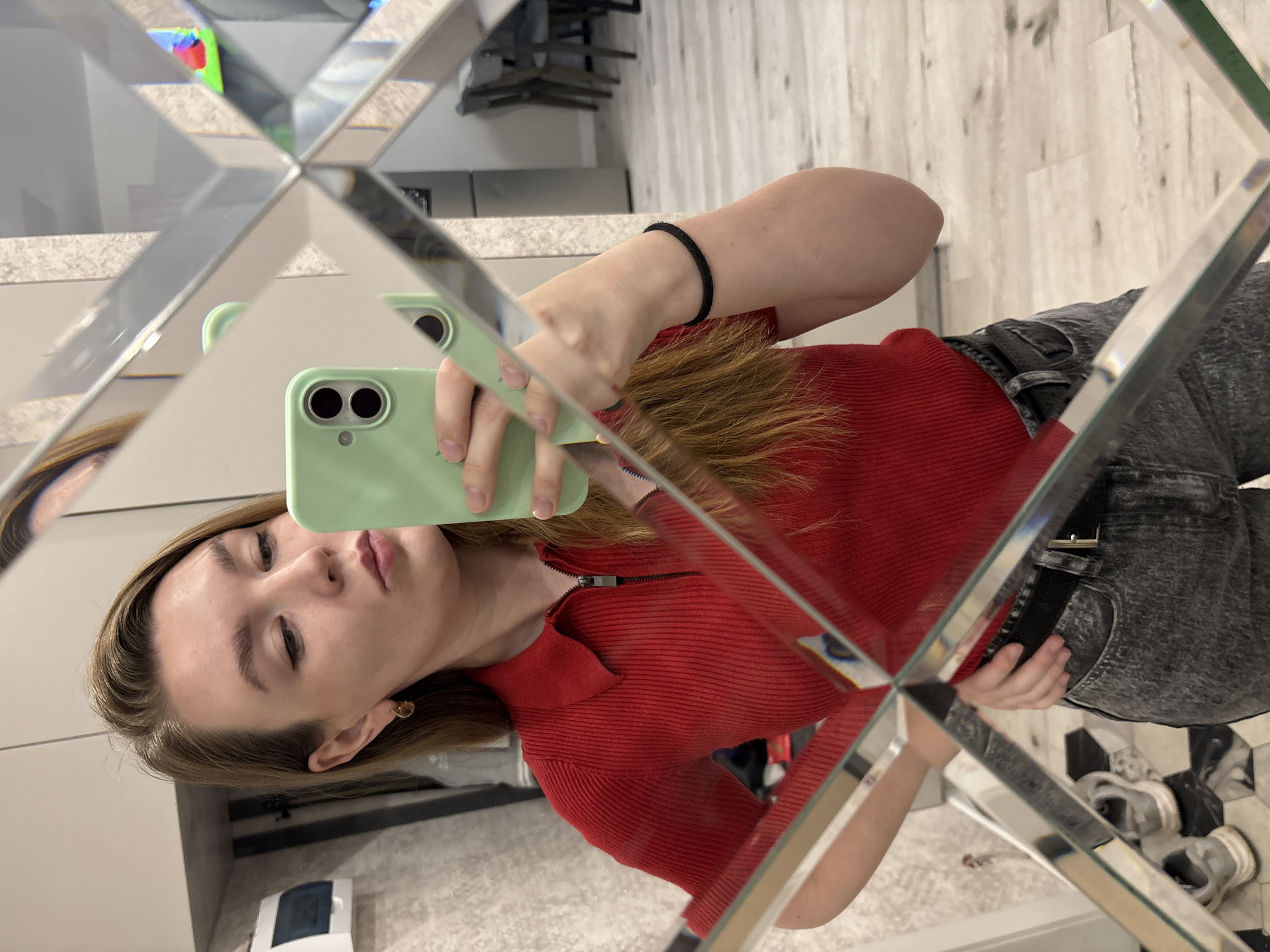A woman is taking a selfie in a mirror. She is wearing a red, zip-up collared shirt, grey jeans with a black belt, and a black hair tie on her wrist. She is holding a light green smartphone. The reflection in the mirror shows part of an interior space, including a wooden floor, a white cabinet, and a clothes rack with hangers. The location is Izhevsk, Russia.