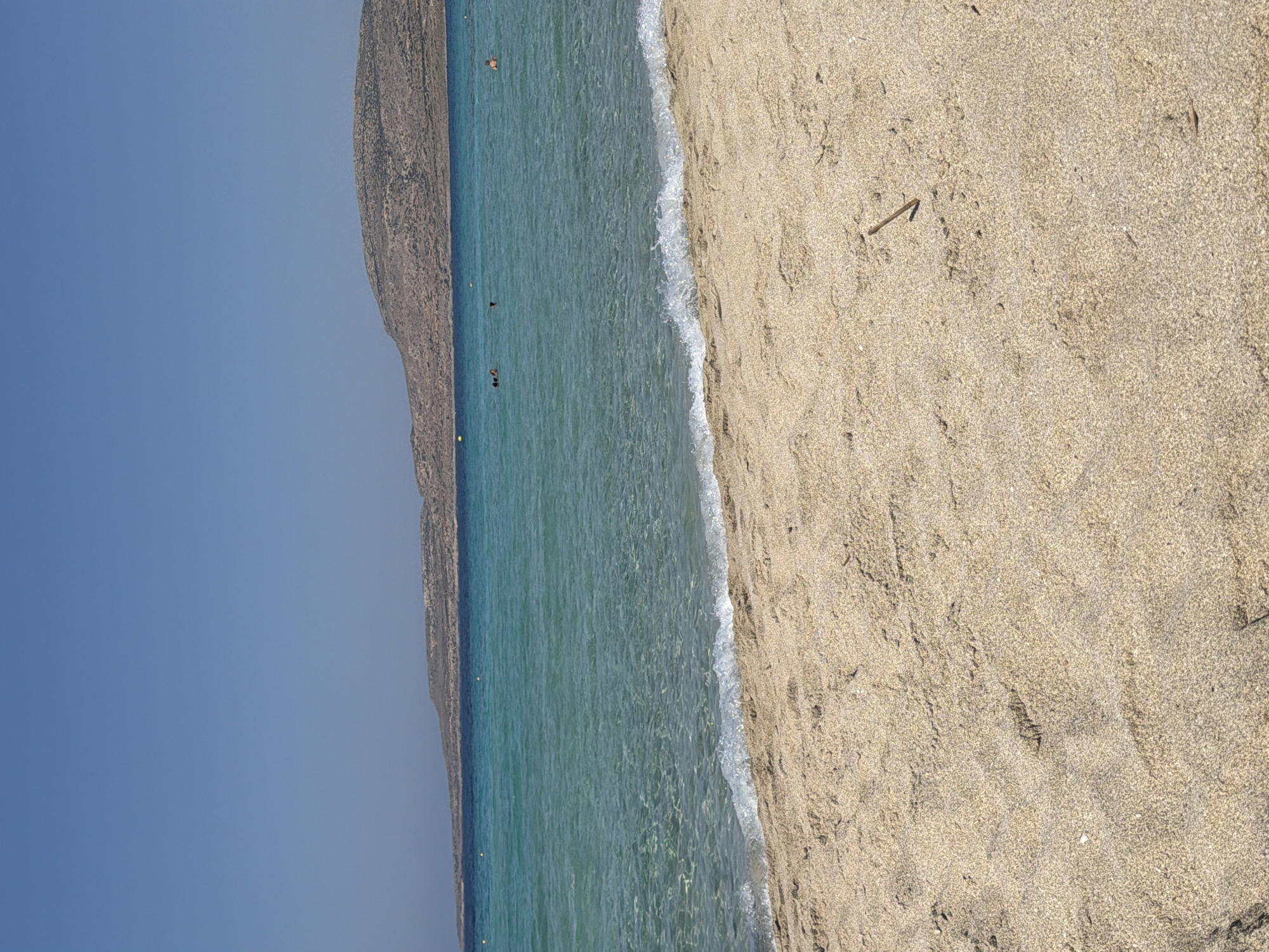 The image depicts a serene beach scene with a clear blue sky overhead. In the foreground, light-colored sand stretches towards the water, showing slight undulations and a small twig. The water is a vibrant turquoise, transparent enough to reveal the sandy bottom near the shore. Gentle waves create a white foam as they reach the beach. In the middle ground, several individuals are visible swimming or wading in the sea. Further in the distance, a body of land with a rocky, arid terrain forms a natural horizon, with a hint of structures on its slopes. The overall impression is a tranquil day at a Grecian beach.