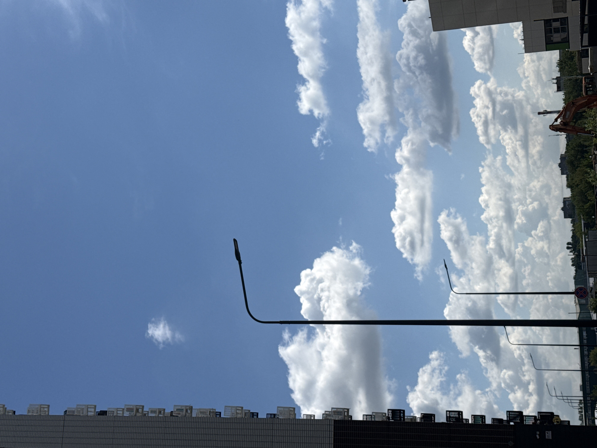 The image depicts an outdoor scene in the city of Lyubertsy, Russia, under a partly cloudy blue sky. In the foreground, a tall, dark-colored building stands on the left side of the frame, characterized by multiple balconies with white railings. A prominent streetlamp with a curved arm is silhouetted against the sky, positioned in the mid-ground to the right of the building. Several other streetlamps are visible further back, receding into the distance. The sky is populated with fluffy, white cumulus clouds. In the background, a line of trees and various structures, including what appears to be a construction site with an orange excavator, are visible. The overall impression is of an urban environment on a bright day.