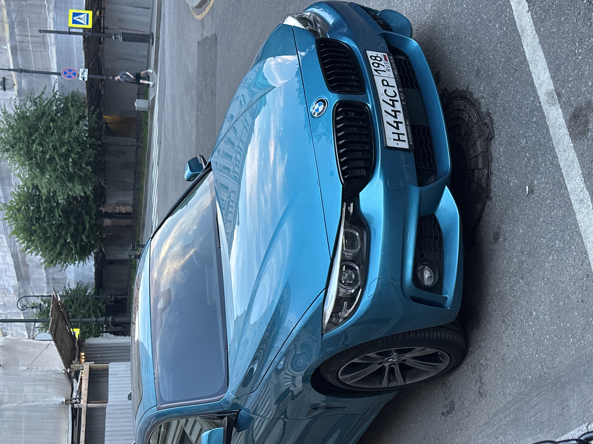 A turquoise BMW 4 Series coupe is parked on a street in Saint Petersburg, Russia. The car has a license plate that reads "H444CP 198 RUS". In the background, scaffolding covers a building, and a person is walking on the sidewalk. The scene is set on a moderately cloudy day.