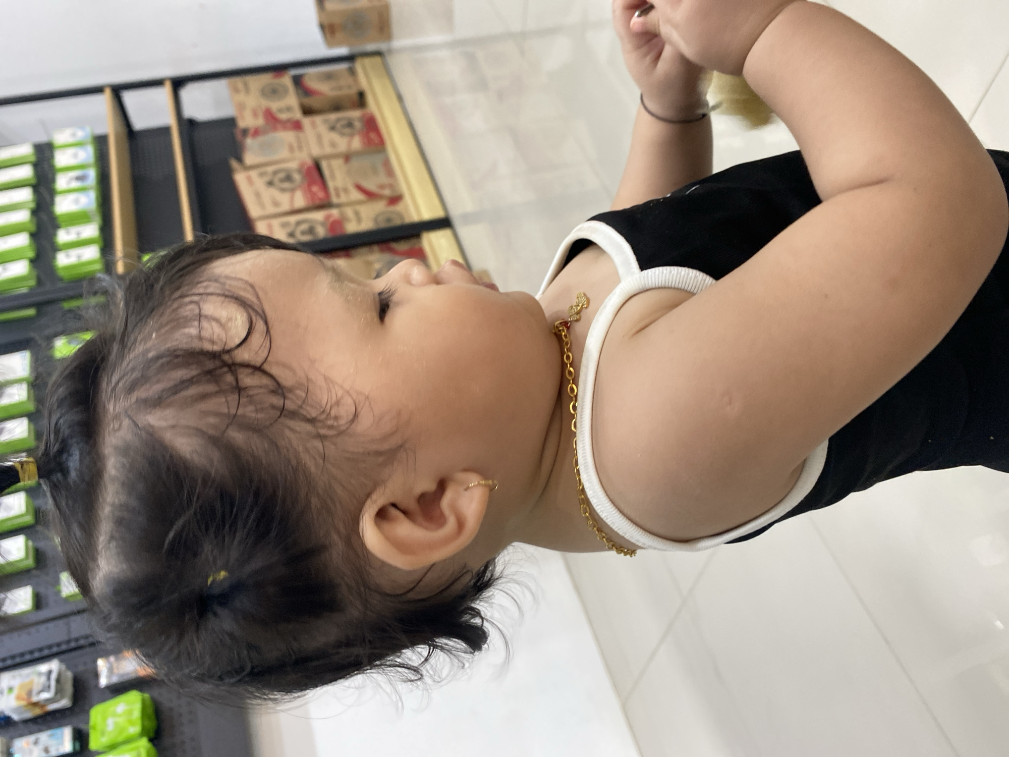 The image shows a young child from the chest up, facing to the right. The child has dark hair styled in two small ponytails on top of their head, with some loose curls around their ears. They are wearing a black tank top with white trim. A thin gold chain necklace with a small, ornate pendant is visible around their neck. A small gold hoop earring is worn in their visible ear. The child's skin is fair. In the background, shelves stocked with various items, possibly electronics or retail goods, are visible. The floor appears to be tiled and reflective. The lighting suggests an indoor setting, likely a retail store. The child's hands are clasped in front of them, holding a yellow object, possibly a toy or cloth. The image is a medium shot.