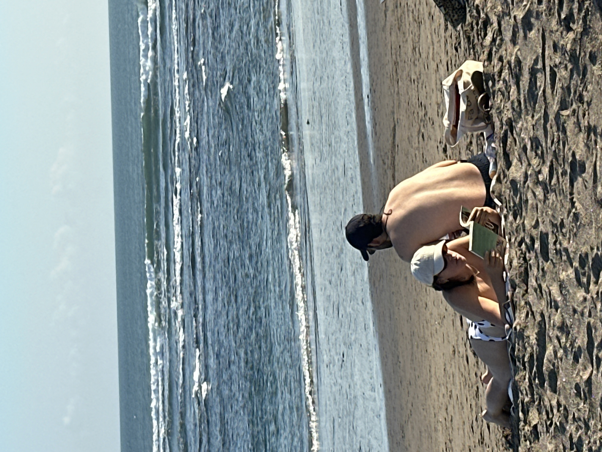 Two people are sitting on a sandy beach in North Kuta, Indonesia, with the ocean in the background. A woman is lying down on her stomach on a towel, wearing a white and black polka dot bikini and a white baseball cap. She is reading a book. A man is sitting behind her, facing away from the camera, wearing a black baseball cap and dark shorts. A canvas bag and a pair of flip-flops are on the sand near the man. The ocean has gentle waves and the sky is clear and blue.