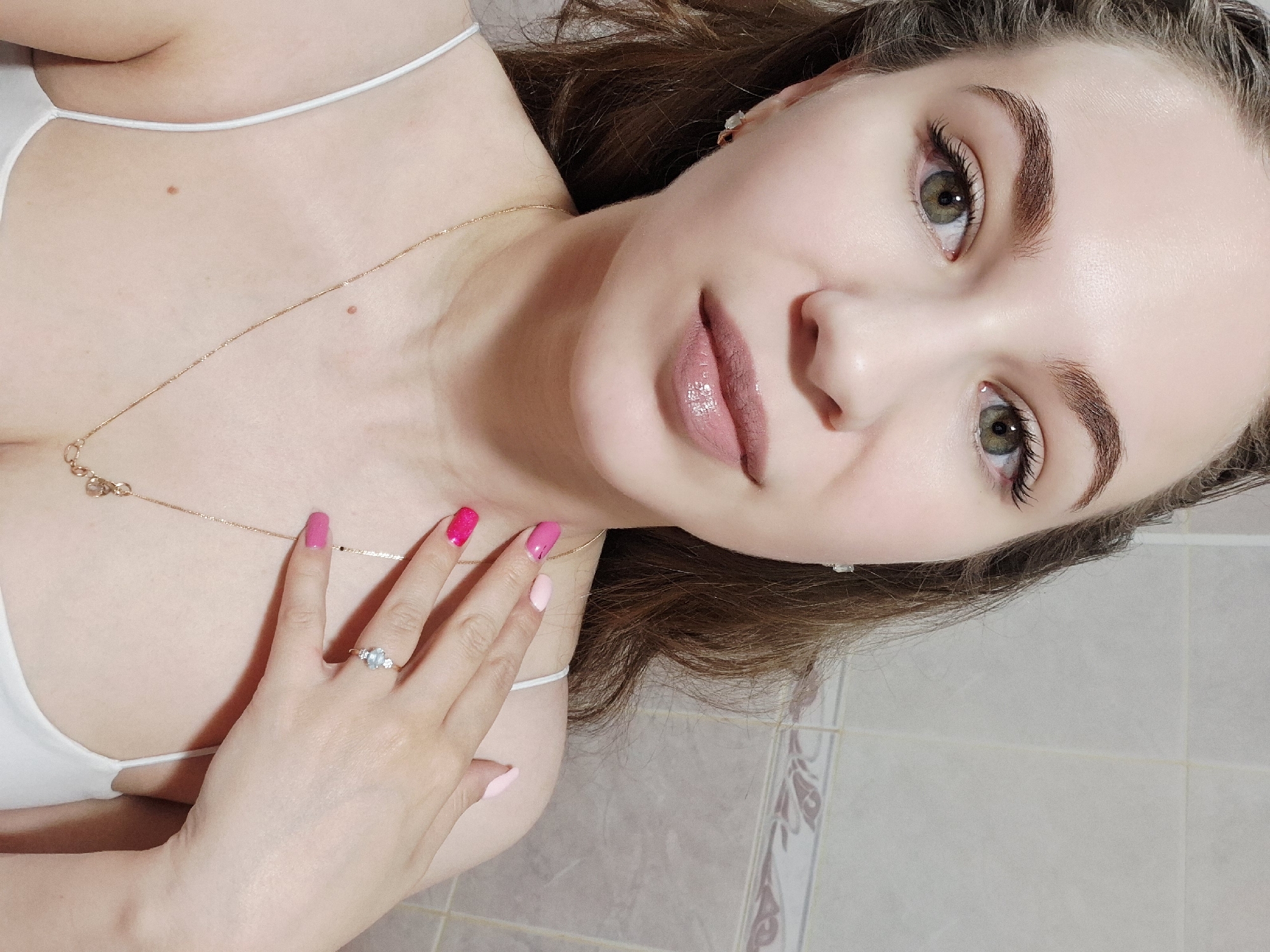 A close-up selfie captures a young woman from a slightly overhead perspective, looking directly at the camera with a calm expression. She has fair skin, green eyes, and well-defined brown eyebrows. Her eyelashes appear long and full, possibly due to extensions or heavy mascara. Her lips are painted with a glossy pink or nude lipstick. She wears a white spaghetti strap top, revealing part of her upper chest where a gold chain necklace with a small pendant rests. Small stud earrings are visible in her ears. Her left hand is placed across her chest, showcasing a manicure with varying shades of pink polish, ranging from fuchsia on her index and middle fingers to lighter pink on her ring and pinky fingers. A ring with a light blue oval stone flanked by two smaller clear stones is worn on her left ring finger. Her brown hair is visible around her head. The background consists of light-colored tiled walls, some featuring a subtle, dark floral or geometric pattern, suggesting an indoor setting like a bathroom or kitchen in Chaikovsky, Russia. There is no visible text.