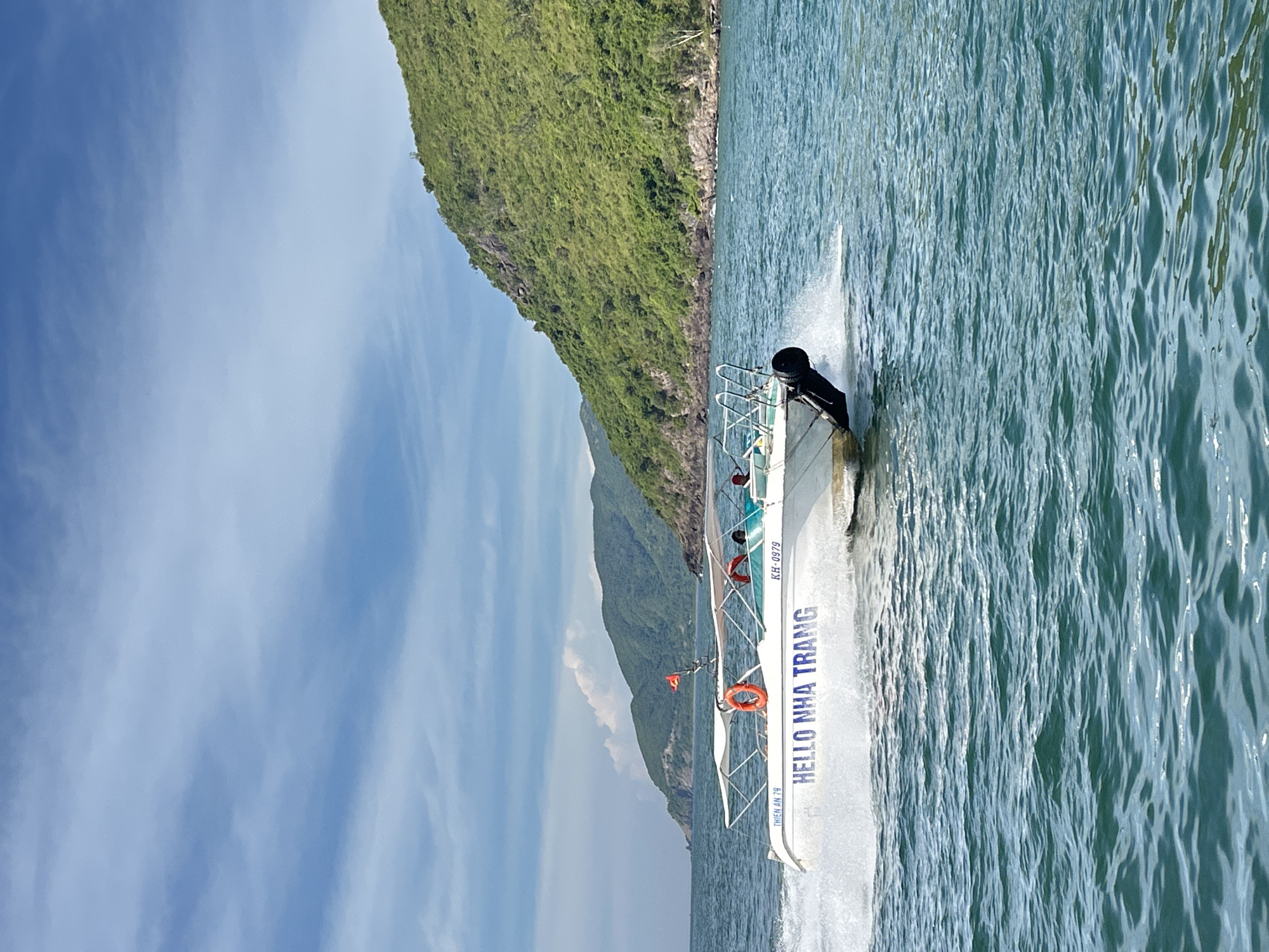 A white motorboat with the words "HELLO NHA TRANG" emblazoned on its side is speeding across choppy blue water. The boat is creating a significant wake as it moves. There are at least two people visible on the boat, one wearing a red life vest. A life preserver ring is visible on the deck. In the background, a lush green, mountainous island rises from the sea. The sky is blue with scattered white clouds. The overall scene depicts a recreational activity on the coast of Nha Trang, Vietnam.