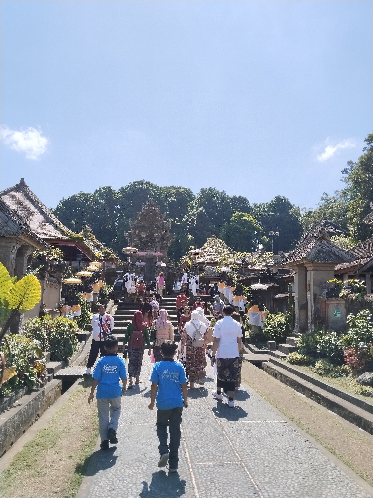 This is a vertical, eye-level, outdoor shot of people walking up a paved pathway towards a temple complex. The sky is clear blue, with a single white cloud. The pathway is made of light-colored cobblestones and is bordered by grassy areas and low stone walls.
On the left side of the pathway, there are several traditional Balinese buildings with ornate, tiled roofs and intricate carvings. Lush green foliage, including large-leafed plants, is visible around these buildings. Several yellow umbrellas are displayed, possibly for shade.
In the center of the image, a wide stone staircase leads up to the main temple structure, which features a multi-tiered roof and weathered stone carvings. More yellow and pink umbrellas are visible on and around the stairs. Many people are ascending the stairs, dressed in a mix of casual wear and traditional Balinese attire, including sarongs.
On the right side of the pathway, more traditional buildings with tiled roofs are visible, interspersed with green vegetation and stone architectural elements.
Several people are the primary subjects in the foreground. Two young boys in blue t-shirts walk away from the camera on the cobblestone path. Behind them, an adult male in a white shirt and sarong walks up the path. Further up the path and on the stairs are various individuals and groups, some in traditional clothing, others in casual attire, all moving towards the temple.
The overall impression is a sunny day at a significant cultural site in Bangli, Indonesia, likely a temple, with a flow of visitors.