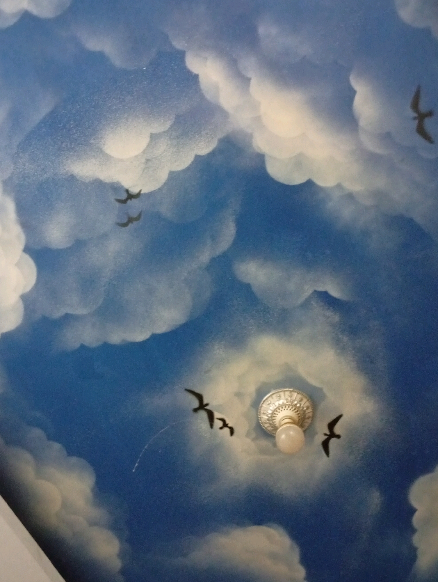 The image depicts a ceiling mural painted to resemble a blue sky with fluffy white clouds. Several silhouettes of birds in flight are scattered across the scene. In the lower center of the image, a light fixture with a round bulb is visible, appearing to be embedded in the ceiling mural. The mural is artfully done, with soft blending of colors to create a sense of depth and realism. The overall impression is one of a serene, open sky. There is no indication of the specific location in Kebondalem, Indonesia, as the image only shows a painted ceiling.
