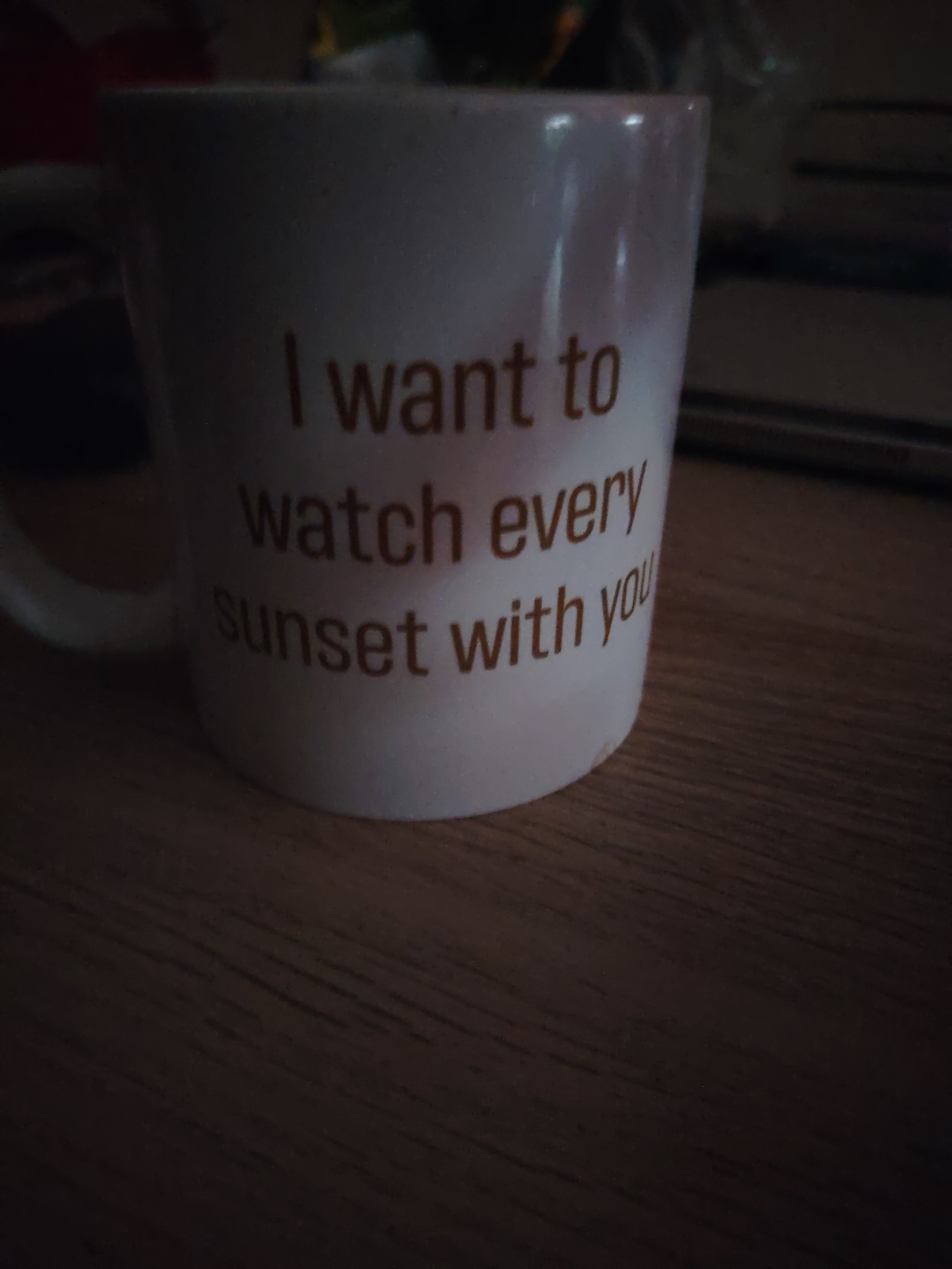 A close-up photograph captures a white ceramic mug resting on a dark wooden surface, likely a table or desk, in an indoor setting within Huntingdonshire, United Kingdom. The lighting is notably dim, suggesting either an evening or night scene, or a poorly lit room.
The mug is the primary subject, with its handle partially visible on the left side. It has a slightly speckled texture. A prominent, sentimental message is inscribed on its side in a clear, brownish-orange sans-serif font, spanning three lines:
"I want to
watch every
sunset with you"
The wooden surface beneath the mug shows distinct grain patterns, running horizontally across the frame. The background is out of focus and very dark, containing vague shapes that could be other household objects or furniture, reinforcing the indoor context. No people or activities are visible; the image solely focuses on the mug and its immediate surroundings. The overall impression is quiet and intimate due to the low light and the heartfelt message on the object.