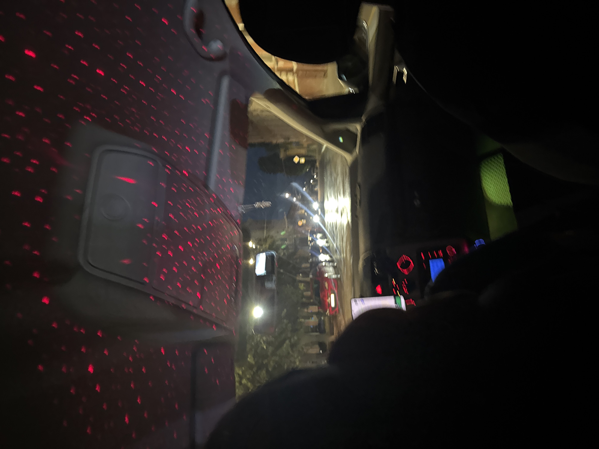 The image shows the interior of a car at night, with red light patterns projected onto the car's ceiling. The view is from the back seat looking forward. Through the windshield, a street scene in Chernivtsi, Ukraine, is visible. Cars are driving on a wet road, and streetlights illuminate the trees and buildings lining the street. The dashboard of the car is partially visible, with a screen displaying information and red illuminated buttons. The scene appears to be a leisurely drive through the city at night.