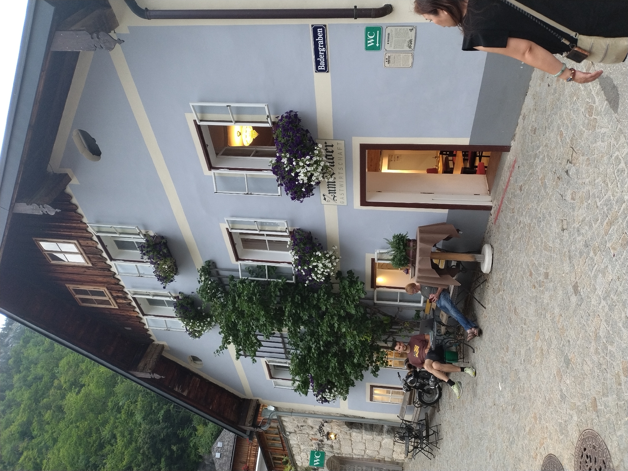 The image shows a street scene in Hallstatt, Austria. A light blue building with a dark wood-shingled roof and wooden trim dominates the frame. The building has multiple windows, some open with flower boxes filled with purple and white flowers. A sign on the building reads "Zum Vader Gastwirtschaft." A woman in a black dress with a tan crossbody bag is walking away from the camera on the right side of the frame. In the foreground, two men are seated at tables outside the building. One man in a brown t-shirt and shorts is sitting with his legs crossed, looking at a laptop. The other man, wearing a gray t-shirt and jeans, is also seated, looking down. A motorcycle is parked behind the men. A WC sign is visible on the left side of the building. Cobblestone pavement covers the street. A lush green hillside is visible in the background.