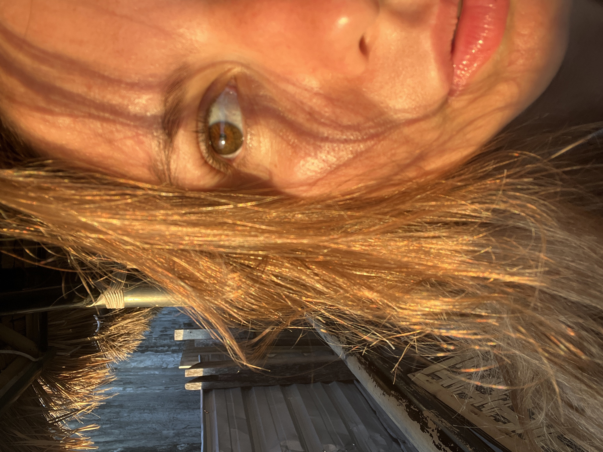 The image is a close-up, low-angle shot of a person's face, likely taken outdoors at sunset. The person has light brown hair, which is illuminated by the golden light of the setting sun, creating a shimmering effect. The light also casts shadows across their face, highlighting their facial features, including their eye and lips. The background is blurred but appears to be a rustic structure with a thatched roof and weathered wooden elements, suggesting a tropical or beachside location. The image's composition and lighting evoke a sense of warmth and natural beauty, possibly capturing a moment of reflection or enjoyment of the sunset.