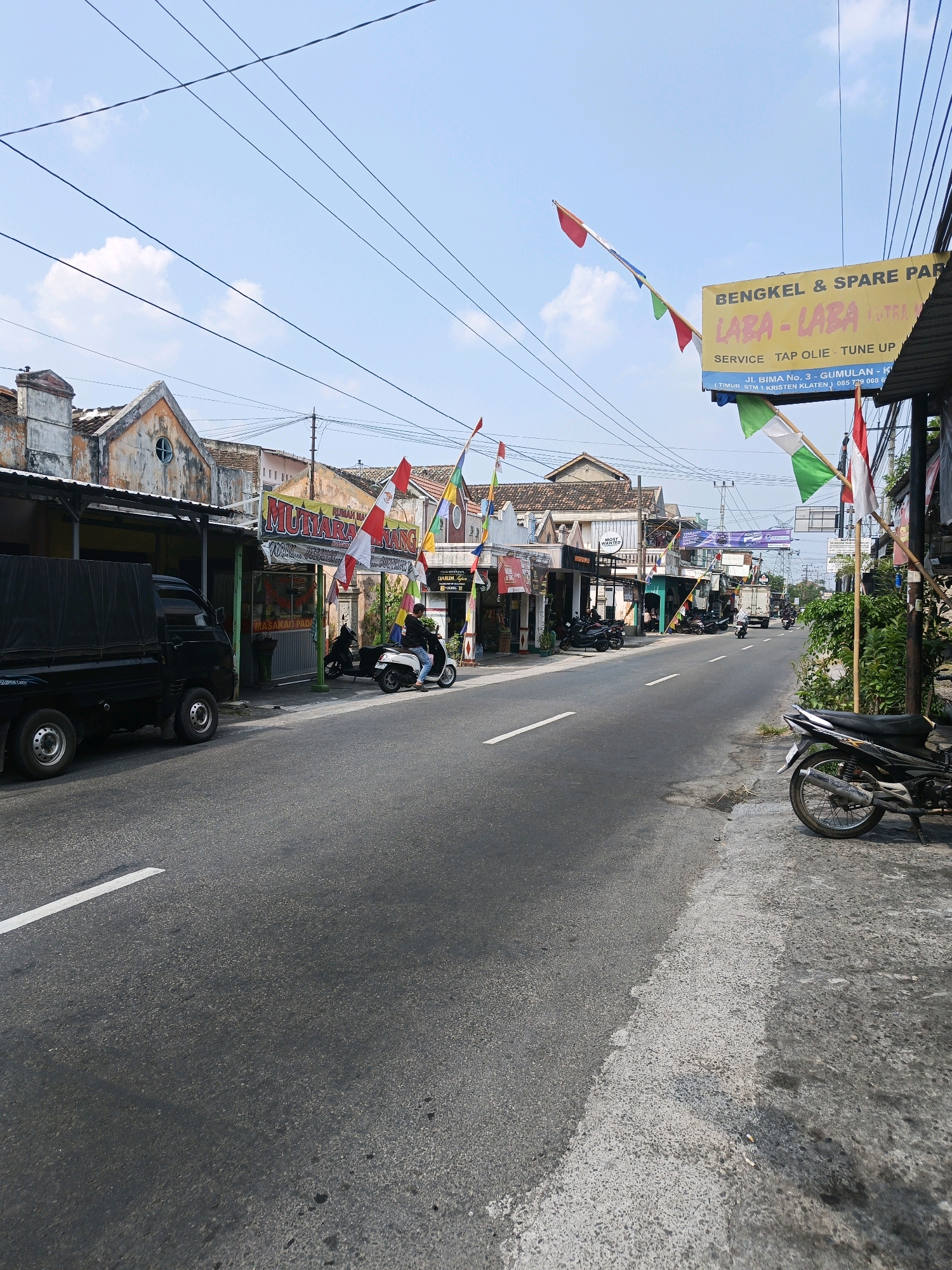 The image depicts a street scene in Karangpakel, Indonesia. The road is paved and has a white dashed line marking the lanes. On the left side of the road, a black pickup truck with a covered bed is parked. Several storefronts line the left side of the street, some with awnings. Red and white flags are strung across the street and attached to poles, some of which are Indonesian flags. A yellow sign with blue lettering is visible, advertising "BENGKEL & SPARE PART" and "LABA-LABA." A person is riding a white scooter on the right side of the road, and another black motorcycle is parked on the right sidewalk. Various trees and vegetation are present along the right side of the street. The sky is blue with some white clouds.