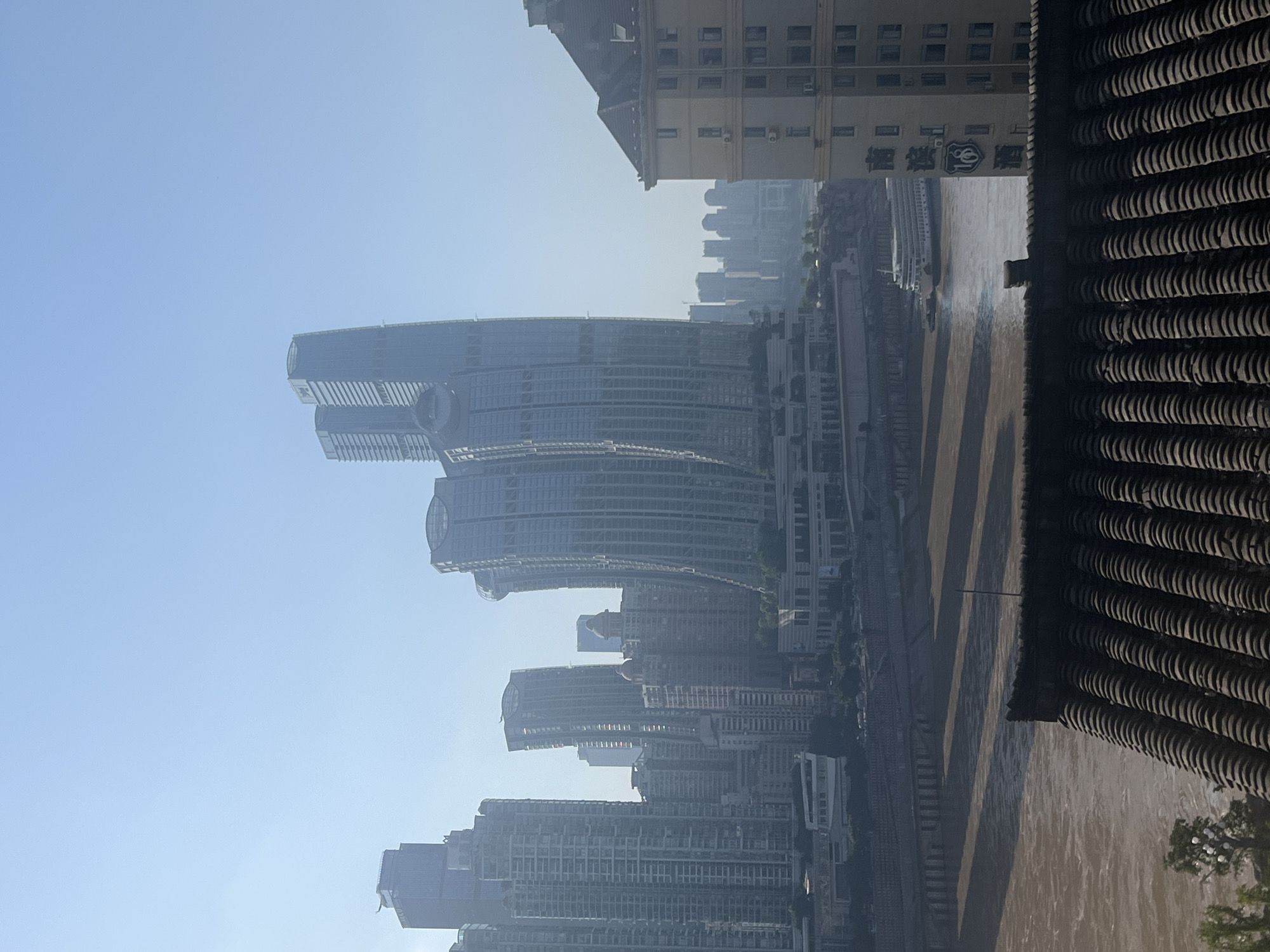 A high-angle, wide shot captures a hazy city skyline in Nan'an District, China, dominated by numerous skyscrapers of varying designs and heights. A wide, muddy river flows in the foreground, with a large white passenger boat docked on its bank. The river is bordered by concrete embankments and a series of tiered buildings. In the immediate foreground, the tiled roof of a traditional Chinese building occupies the bottom portion of the frame, casting dark shadows on the river. The sky is a pale, hazy blue. The scene appears to be captured during the daytime, likely in the morning or late afternoon, given the soft lighting and haze. No people are visible in the frame.