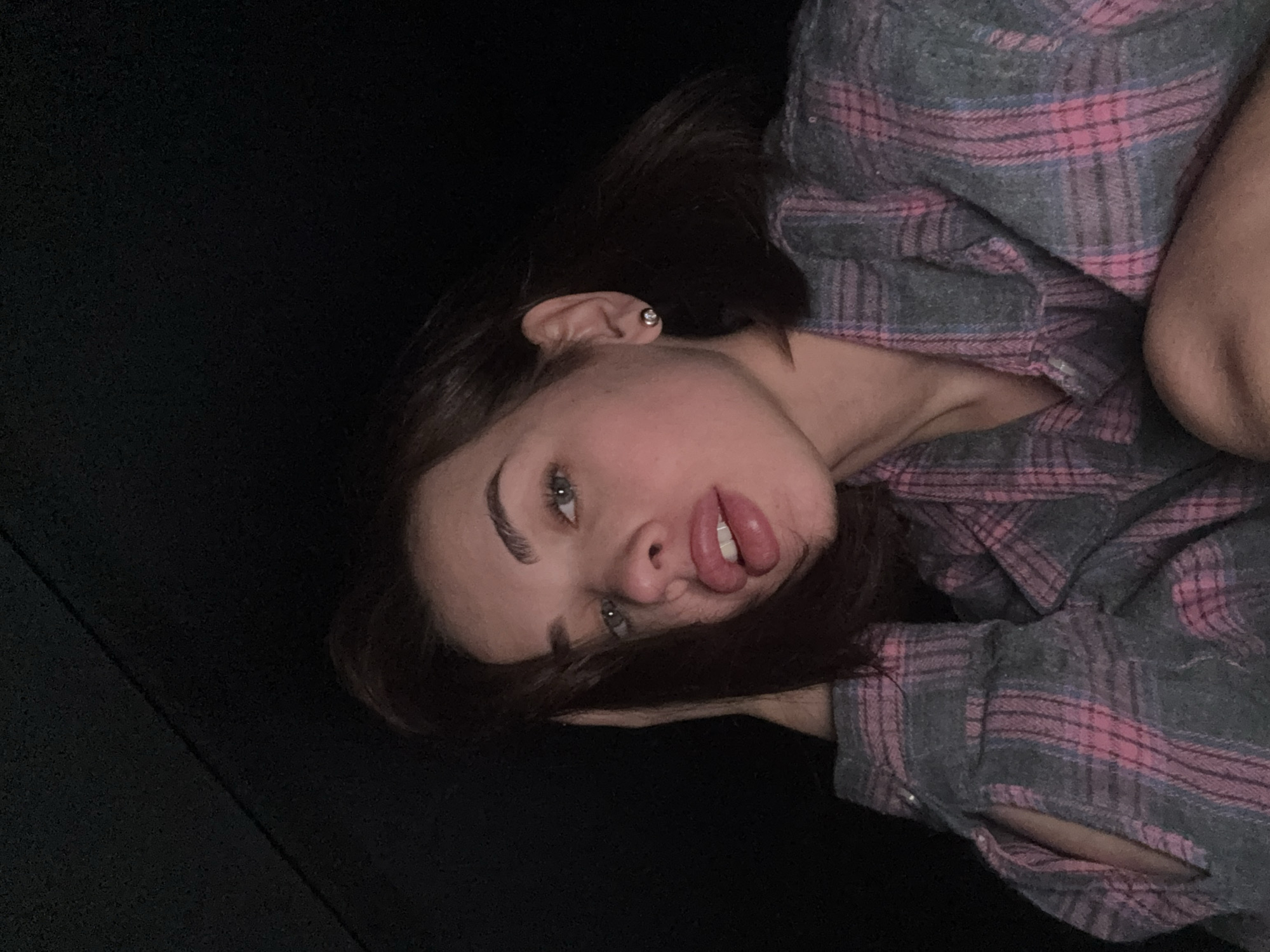 A young woman lies on a dark surface, possibly a couch or bed. She is angled towards the camera with her head tilted. She wears a gray and pink plaid flannel shirt and a small earring is visible in her left ear. The background is dark and nondescript. The location is unspecified as there is no evidence of the city of Лобаново, Russia in the image.