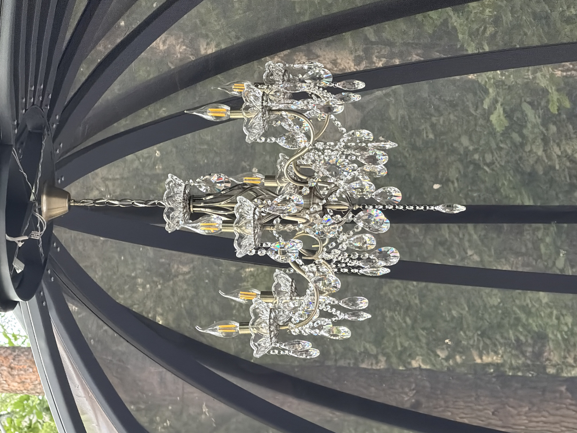 The image shows a crystal chandelier hanging inside a gazebo or similar outdoor structure with a black mesh roof and frame. The chandelier is ornate, with multiple crystal droplets and candle-shaped LED bulbs. The background is blurred, showing trees and greenery, suggesting an outdoor setting. There are no people or animals visible in the image. The location is described as Rakitnitsky Sel'ski Sovet, Belarus, though this specific detail cannot be verified from the image alone.