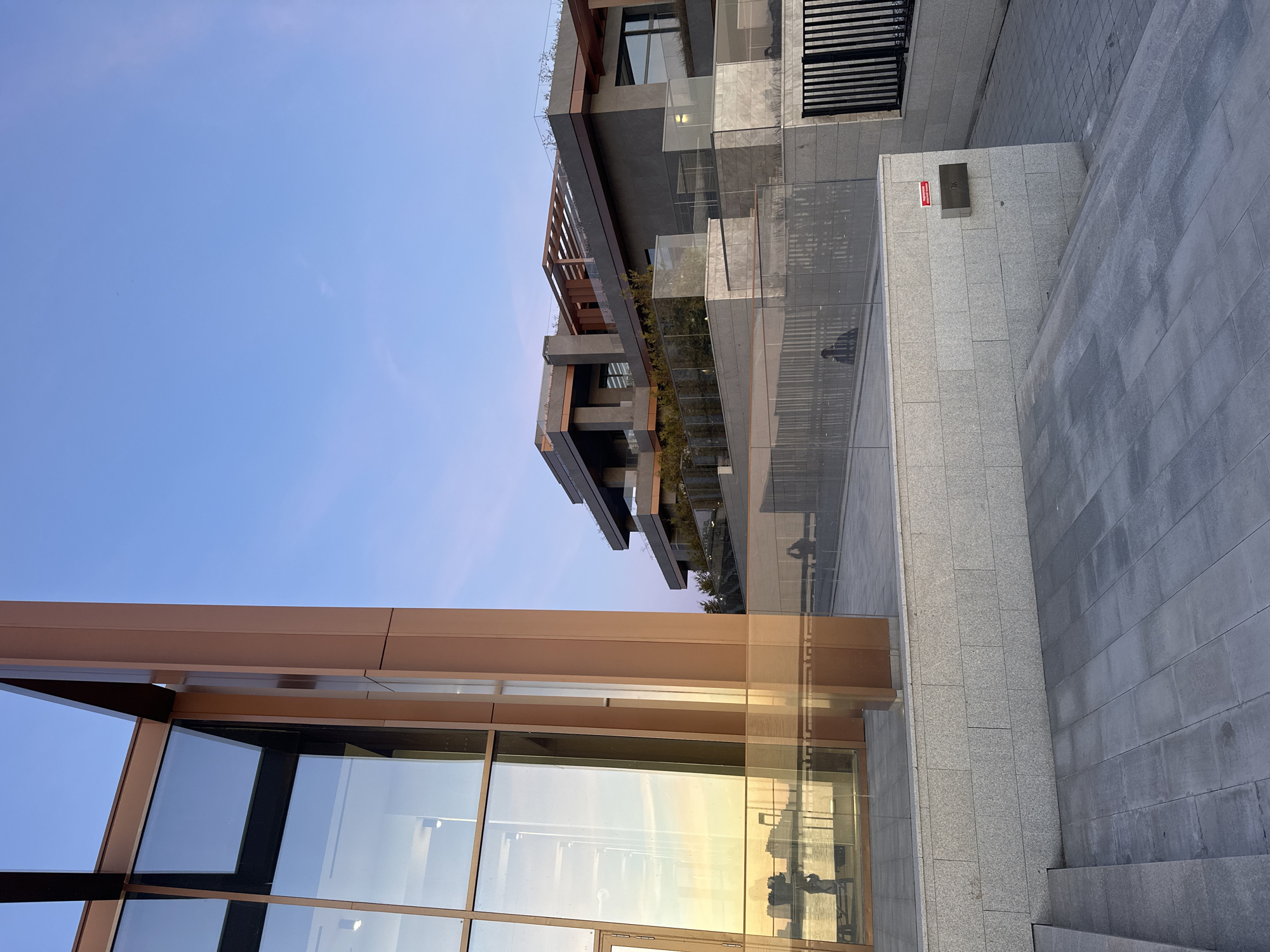 The image displays a modern urban architectural scene, captured during what appears to be the late afternoon or early evening due to the quality of light. The sky, visible in the upper left, is a clear, bright blue with no clouds.
In the foreground, on the left side, a contemporary building structure is prominent, characterized by large glass panels set within broad bronze-colored framing. These glass surfaces strongly reflect a warm, golden light, indicative of a low sun angle, and reveal the faint, distorted silhouette of a person standing.
Extending to the right of this structure is a paved outdoor area. It features a raised section with light grey square tiles, and further right, steps made of darker grey rectangular pavers descend. On a light grey wall within this paved area, a small red rectangular sign is mounted, clearly displaying a white crossed-out cigarette graphic, signifying "NO SMOKING." Below this sign is a dark metal box with the letters "TW" or "TVC" faintly inscribed.
In the background, a multi-story modern building rises, its facade composed of dark grey cladding, extensive glass balconies with railings, and sections adorned with greenery. Architectural details include wooden or bronze-colored slatted elements and beams. A subtle human silhouette can also be seen reflected in one of the glass railings of this building.
No direct activity or event is depicted, but the reflections of individuals suggest a public space. The scene conveys a sense of contemporary urban design, blending materials like glass, metal, and stone with touches of nature, all bathed in the gentle, golden hues of the setting sun. The location is Kazan, Russia.