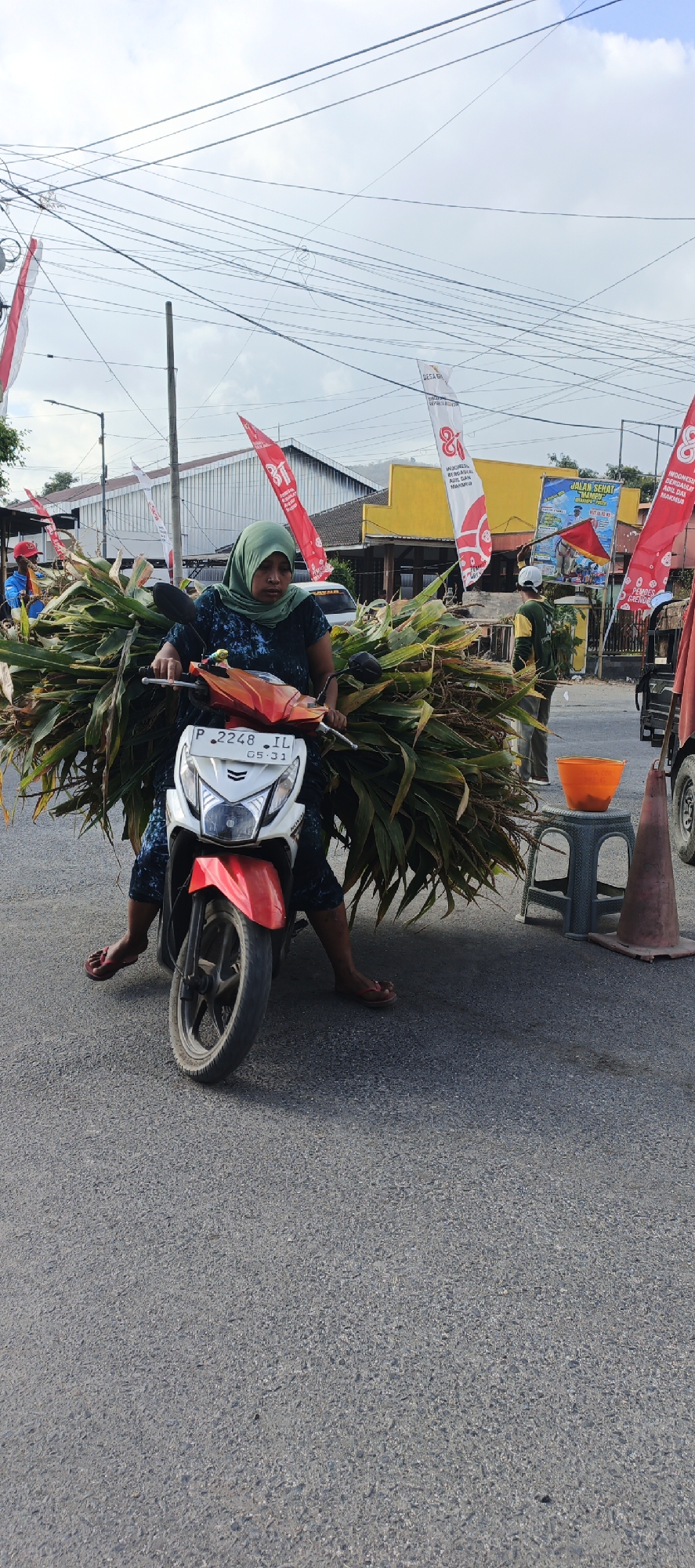 A woman wearing a green hijab and a dark blue patterned dress is seated on a white and red scooter. She is transporting a large bundle of corn stalks that are tied together and extend out from behind her on both sides. Her feet, clad in red sandals, are resting on the ground. The scooter's license plate reads "P 2248 IL 05-31". The scene appears to be on a paved road, with buildings and banners in the background. The sky is overcast. A small orange bucket is visible on a green stool to the right of the scooter, next to an orange traffic cone. There are also red and white banners with writing in Indonesian displayed on poles. The overall setting suggests a rural or semi-urban area in Grenden, Indonesia, likely during daylight.