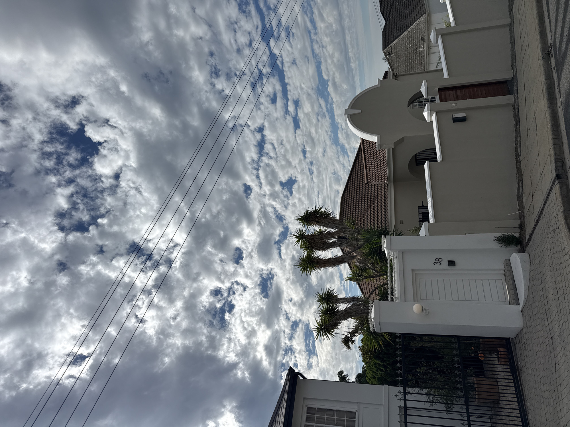 A street in Cape Town, South Africa, shows a white building with a black gate in the foreground, and a tiled walkway. The building has the number "38" on the wall above the door. In the background, there are other white buildings with brown tile roofs and palm trees in front. Overhead, there are power lines against a partly cloudy sky with blue patches. The image is oriented vertically.