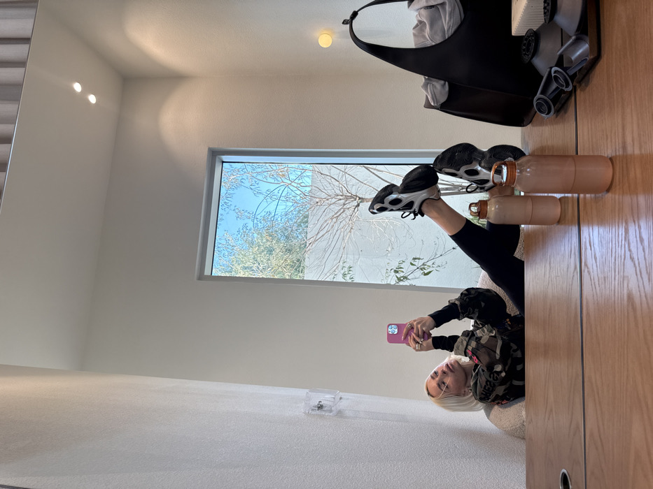 A woman reclines on a couch with legs stretched upward, wearing black leggings and athletic shoes, taking a selfie with a pink phone. A window behind her displays a clear sky and leafy trees. On the right, two water bottles sit next to the woman's feet on a wooden surface, along with a black purse and gray cylindrical objects. The room features minimalist white walls and recessed lighting. A small, clear container sits on a ledge near the bottom left corner.