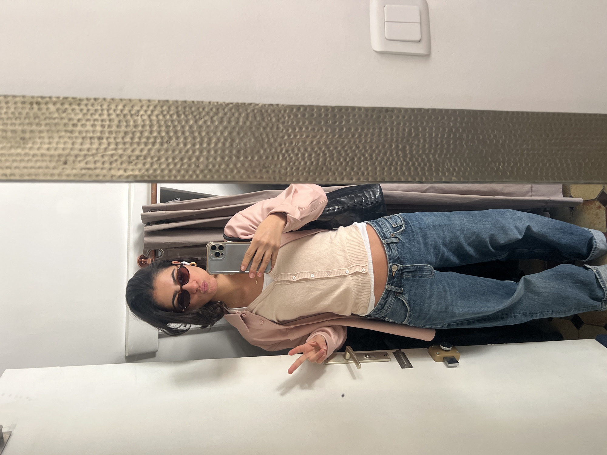 A young woman captures a mirror selfie in an indoor setting. She is reflected standing upright, dressed casually in dark sunglasses, a light pink cardigan, a lighter-colored button-up top, a white undershirt, and blue jeans that are cuffed at the ankles. She holds a smartphone to take the picture, pouting her lips and making a peace sign with her right hand. A black handbag is visible on her left side.
The background reflected in the mirror shows a white wall with a white light switch positioned towards the top right. Below this, a textured, horizontal panel with a dimpled or hammered metallic appearance stretches across the mid-section. Behind the woman, glimpses of a draped curtain or fabric are visible. The lower part of the reflection reveals a white surface, possibly a counter or door, featuring a visible door latch mechanism. The reflected floor appears to have patterned tiles. The lighting suggests daytime. There is no visible text. The overall mood is playful and casual.