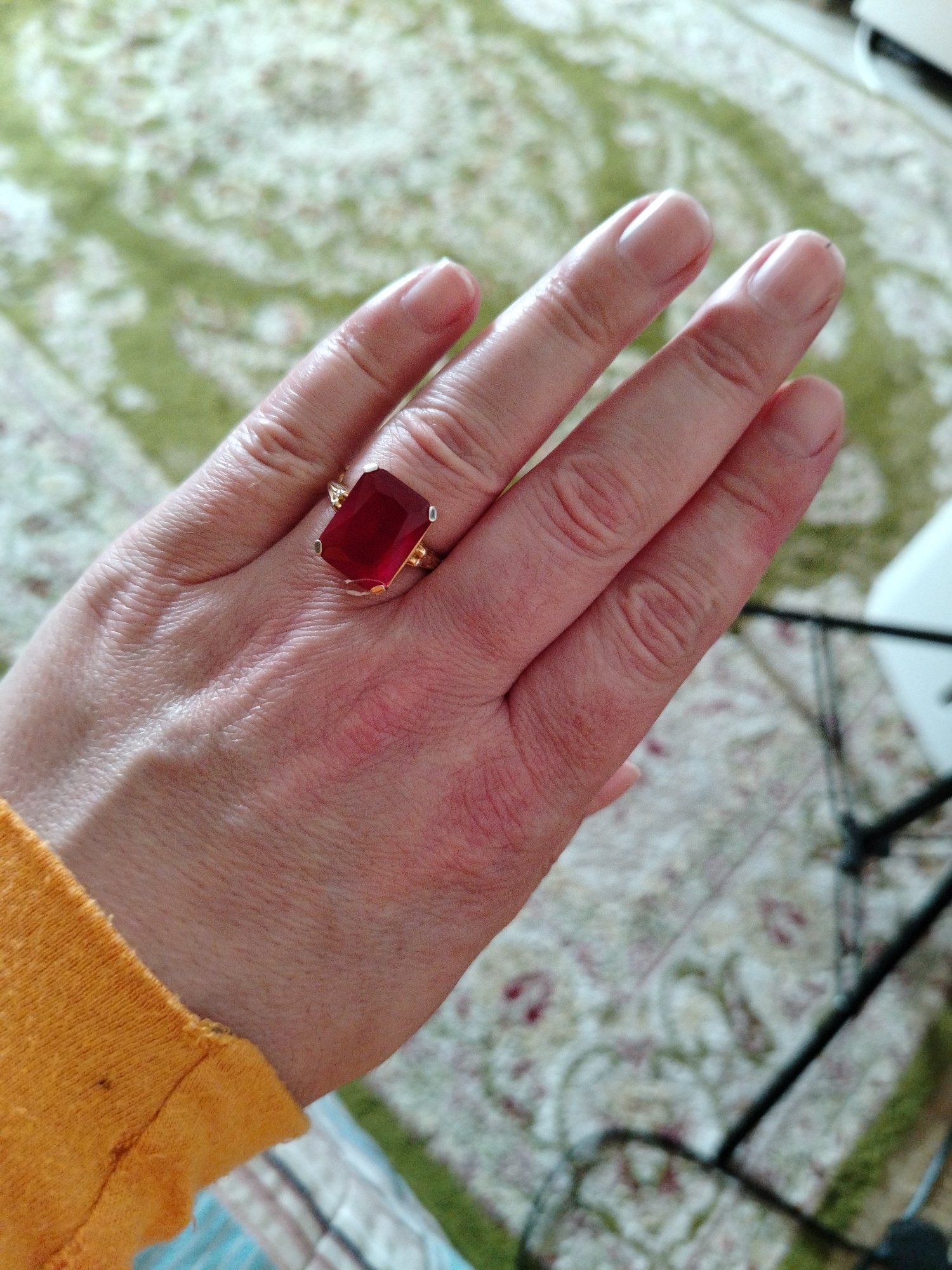 A hand with a large ruby ring is displayed. The ring is gold with a large rectangular ruby. The hand is light-skinned and appears to be wearing an orange sleeve. The background is a patterned carpet and a black metal object. The image's location or whether it was taken in the city of Al Mas'oudi, United Arab Emirates, cannot be determined.