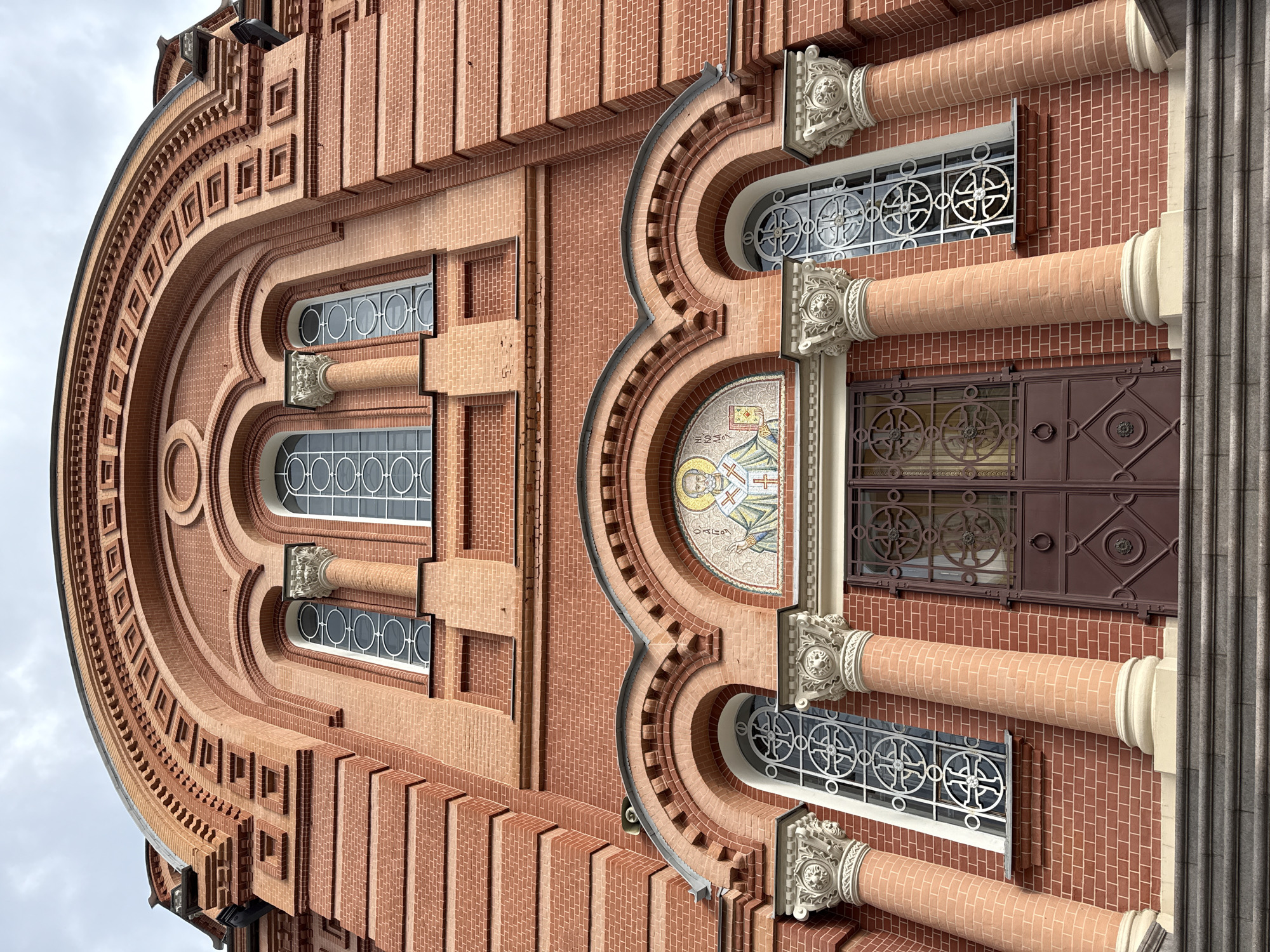 This is an exterior architectural photograph of a building, potentially a church or cathedral. The building is primarily constructed of reddish-brown brick. The facade features an arch-shaped design element at the top, with detailed brickwork including recessed squares. Below the arch, there are rectangular windows with white frames. The windows contain circular designs within them.
The middle section has an arched element with an icon or mosaic design in the center. Flanking this central design are more rectangular windows with similar circular patterns. Column-like structures made of brick and topped with ornate white capitals support the arches.
On the right side, there is a door, which is also located between two windows and brick columns with capitals. The door is dark brown with metalwork featuring circular patterns. A gray stone column is present on the right edge of the image.