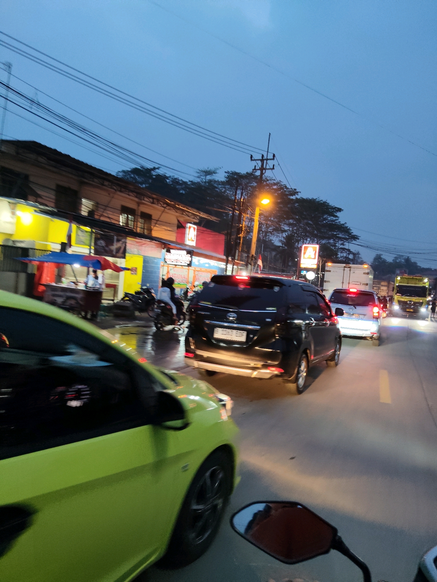 The image depicts a street scene in Kaduagung Tengah, Indonesia, at dusk. A bright yellow car, partially visible in the foreground, is in motion, followed by a black SUV. Further ahead, a silver car and a white truck are also on the road. The street is wet, suggesting recent rain, and lined with shops and stalls on the left side, some of which are illuminated by yellow and red lights. Power lines are strung across the scene, and a street lamp is lit. Several motorcycles are parked and one is being ridden, with two individuals on it. The sky is a clear, pale blue, indicative of twilight.